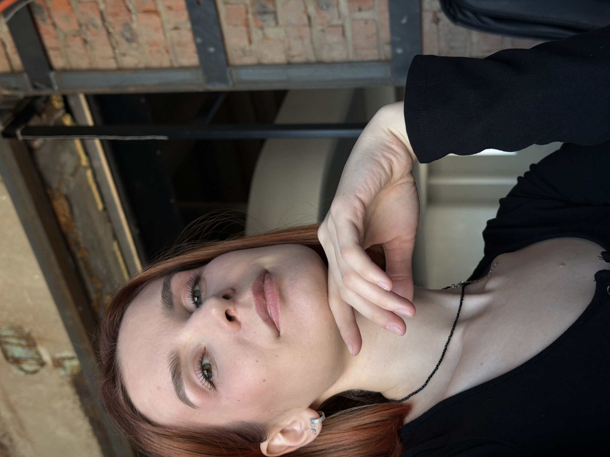 A portrait of a young woman with light skin, auburn hair, and green eyes. She wears a black top and a delicate silver necklace. Her right hand is gently placed near her face, and she has an earring in her left ear. The background includes brickwork and industrial-looking metal beams, possibly indicating an urban location such as an interior space in Chaikovsky, Russia.