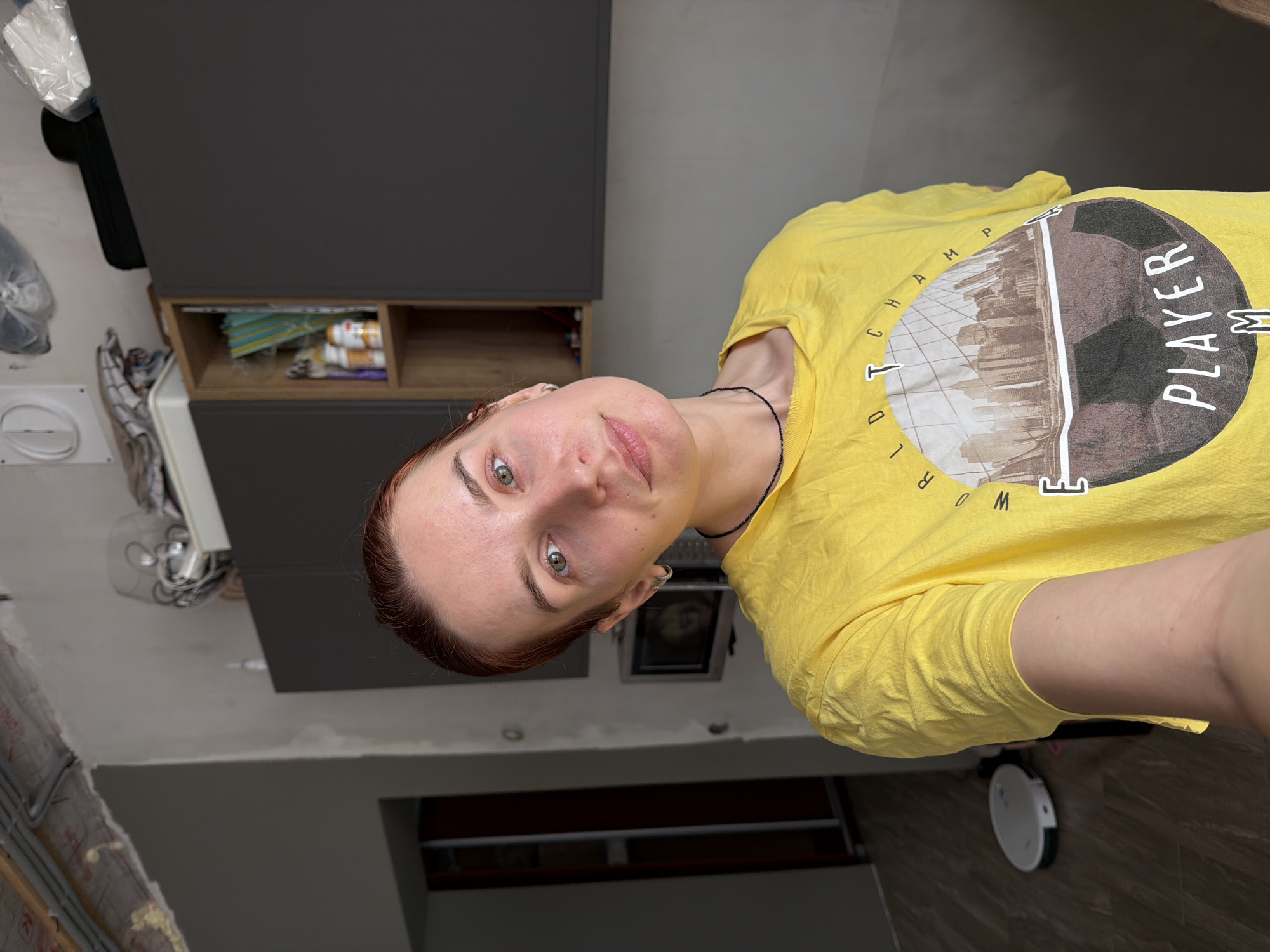 A woman wearing a yellow t-shirt with a circular graphic that includes the words "WORLD CHAMP" and "PLAYER" is in an interior setting, likely a kitchen. The setting includes modern gray kitchen cabinets, a visible collection of items in a cabinet above a counter, exposed wiring and unfinished wall construction, and a white robotic vacuum cleaner on a brown-toned tiled floor. The location is possibly in Chaikovsky, Russia, though there is no visual confirmation of this.
