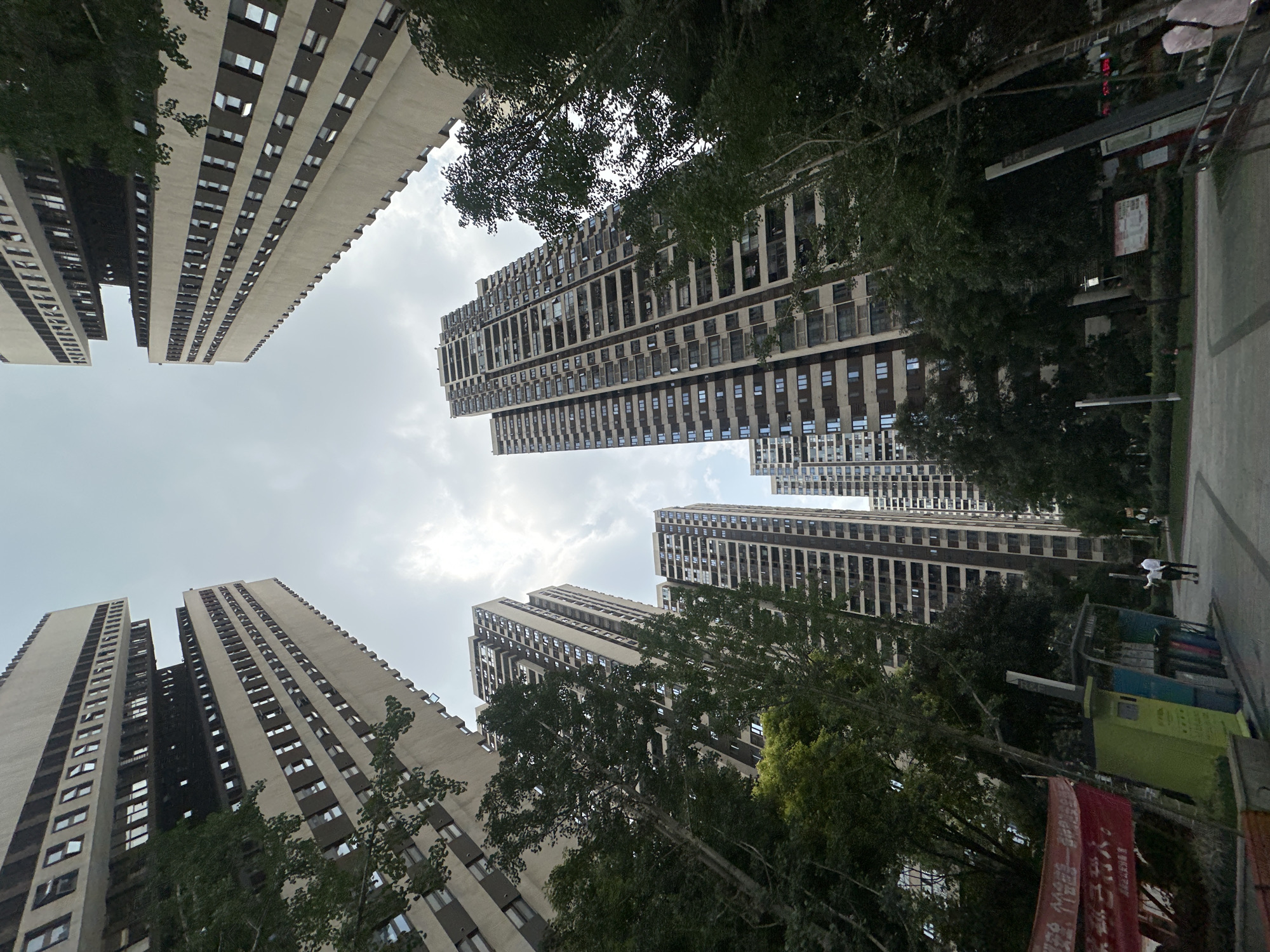 The image shows a low-angle view of tall apartment buildings in the Chongqing High-tech Zone, China. Several buildings of similar design rise into a cloudy sky. Trees with green foliage are interspersed among the buildings, partially obscuring the lower floors and creating a sense of depth. On the right side of the image, the corner of a building and part of a street or sidewalk are visible, along with some infrastructure, possibly bins or street furniture. Two people, dressed in white, are walking down the street.