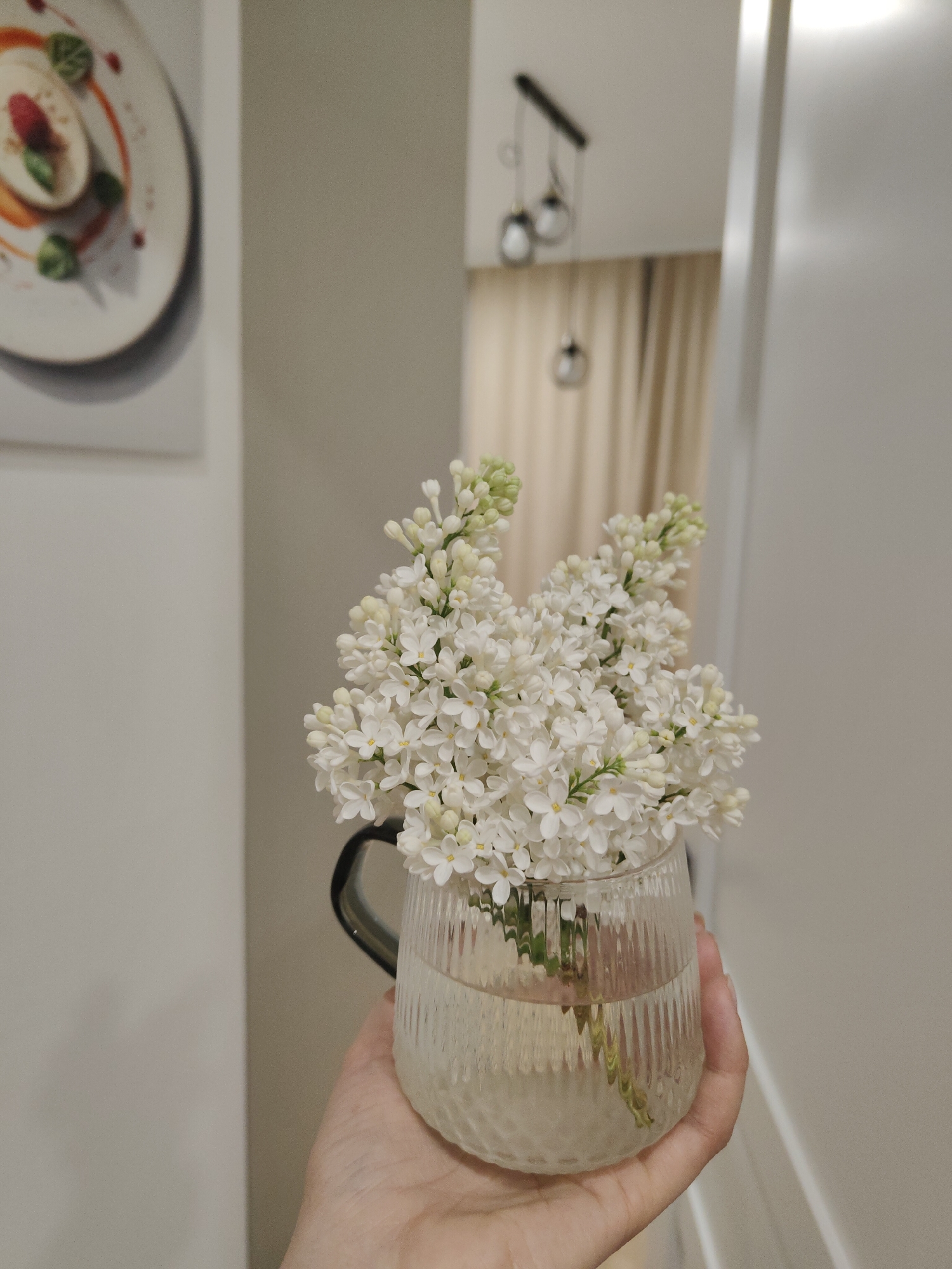 A hand holds a transparent glass mug filled with water and white lilac flowers. A white wall with a framed picture of a dessert is to the left of the mug. In the background, a hallway with light tan curtains and a modern light fixture with round bulbs hanging from the ceiling is visible. The location is an interior setting in an unidentified city.