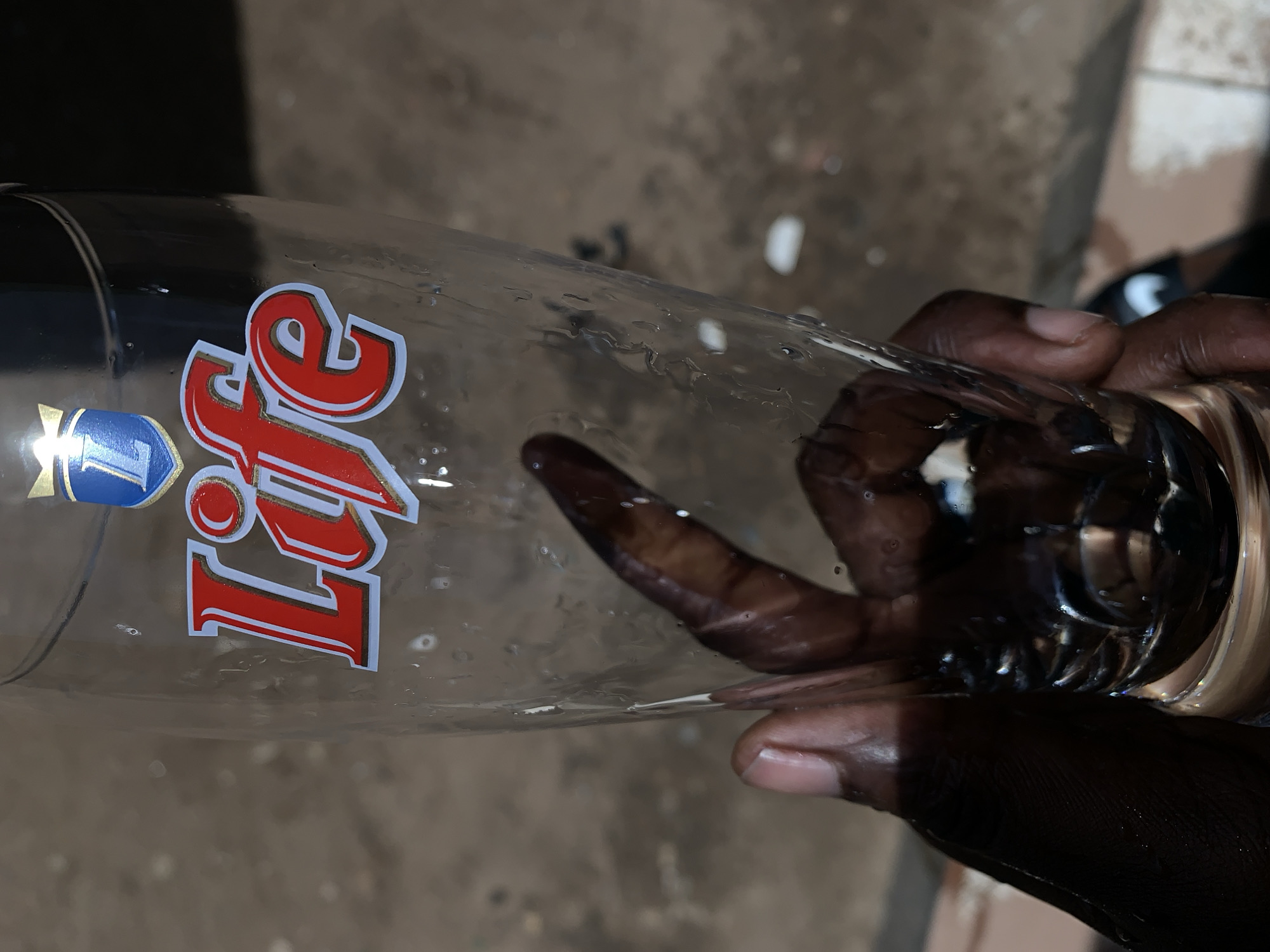 The image shows a close-up view of a person's hands holding a clear glass. The hands have dark skin and are positioned around the lower and middle sections of the glass. The glass is adorned with a prominent label that features the word "Life" in bold red letters with a gold outline. To the left of "Life" is a blue and gold emblem displaying a stylized "L".
Numerous water droplets are visible on the outer surface of the glass, suggesting it might have contained a cold drink or been recently washed. The glass appears to be mostly empty. The background is out of focus, depicting a muted, earthy-toned surface, possibly concrete or ground, consistent with an outdoor or semi-outdoor environment. The scene is illuminated by natural light, indicative of daytime. The context provided places this scene at the New Stadium in Nigeria.