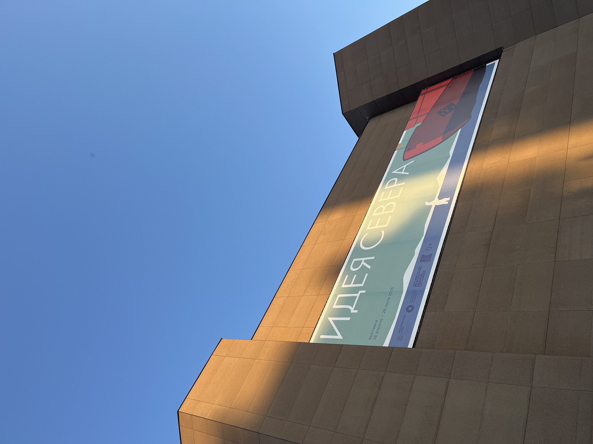 The image shows a building with a large banner displaying Cyrillic text. The text is "ИДЕЯ СЕВЕРА" and smaller text below it states dates "18 апреля - 26 июля 2026." The building's facade is covered in rectangular tiles. A portion of the building is illuminated by sunlight, creating a stark contrast with the shadowed areas. Above the building is a clear blue sky. The location of the building is Unknown, Unknown.