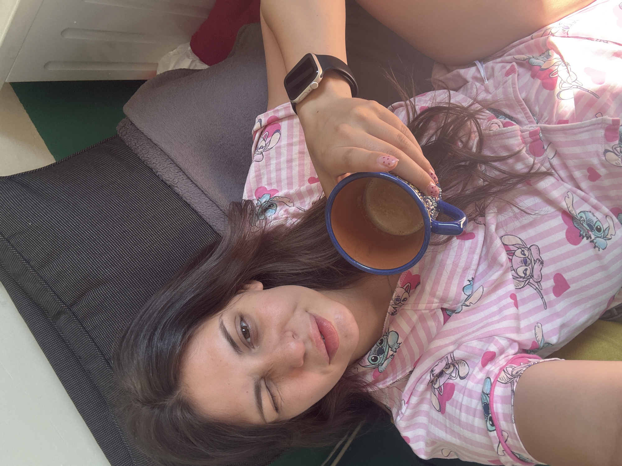 A woman with dark brown hair is lying down, holding a blue mug with a beverage in her right hand. She is wearing pink and white striped pajamas with Stitch cartoon characters and pink hearts printed on them. She is winking, and her lips are slightly parted. On her left wrist, she is wearing a black smartwatch. The background shows a dark grey cushion and a light-colored wall. The image is a selfie, taken from a high angle. The scene appears to be indoors, possibly in a bedroom or living room. There is no identifiable landmark or specific location within the city of Plovdiv, Bulgaria.