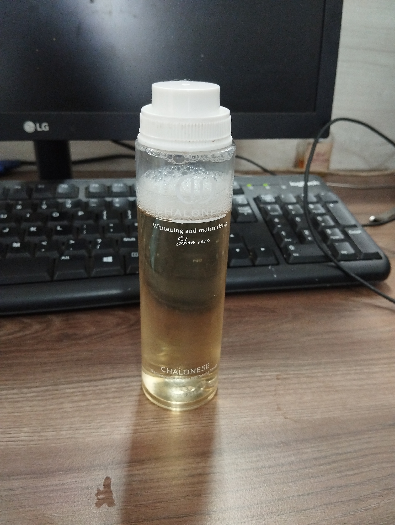 A clear plastic bottle with a white cap contains a pale yellow liquid with foam at the top. The bottle has a label with white text that reads "CHALONESE" and below it "Whitening and moisturizing" and "Skin care". The bottle is positioned on a wooden surface. In the background, a black computer keyboard is visible, and behind that, a black computer monitor with the LG logo on the lower left. The scene appears to be an indoor setting, likely a desk area. There is no specific indication of its location within Kelapa Dua, Indonesia.
