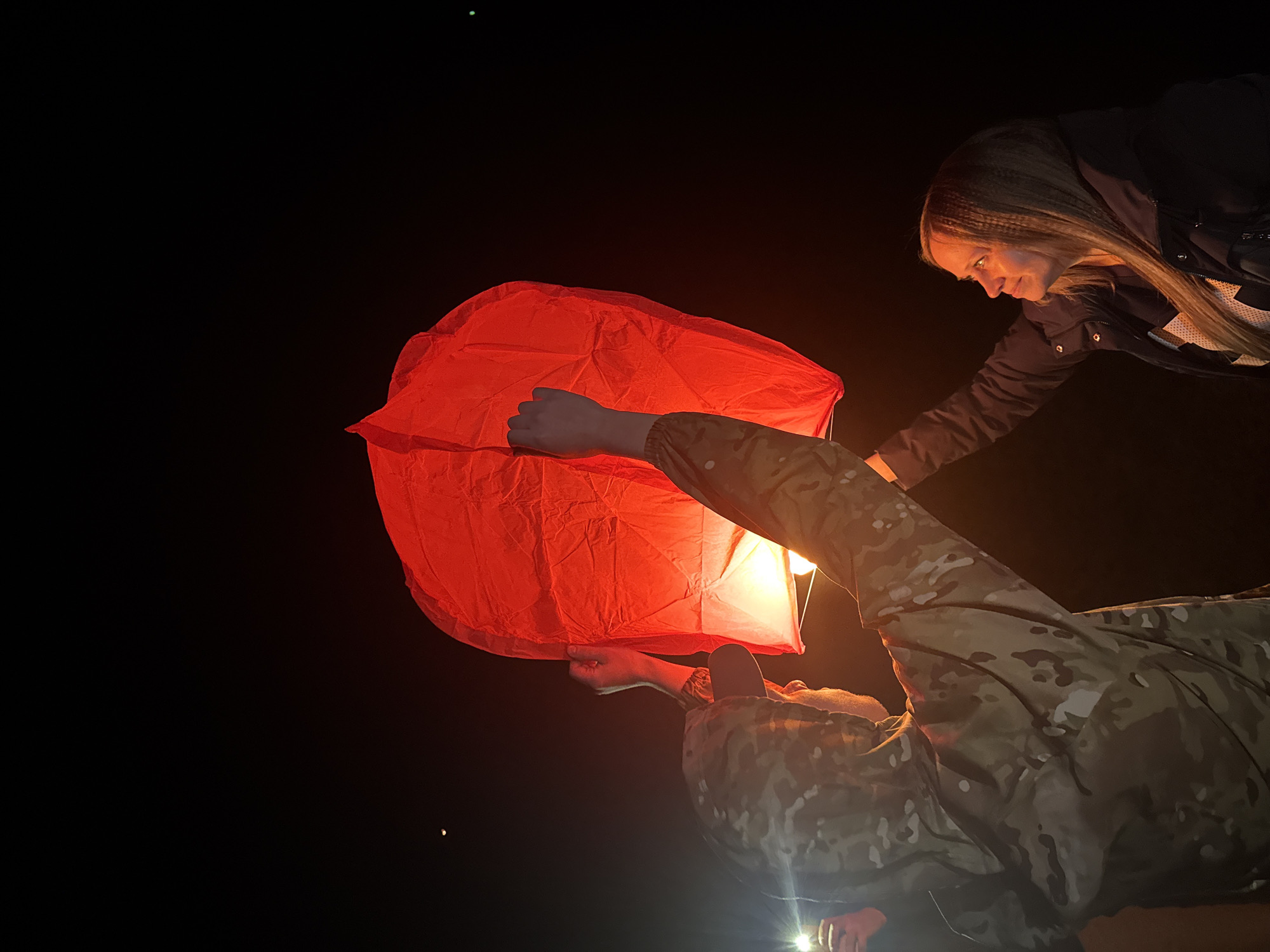 The image captures two individuals at night, preparing to launch a red sky lantern outdoors. The setting is predominantly dark, with the only significant illumination coming from the glowing lantern and a bright light source near the lower person's waist.
In the foreground, a person wearing a camouflage-patterned jacket is holding the large, red paper lantern from below, appearing to look up into it. Their face is mostly obscured. A strong, possibly artificial, light emanates from below this person, casting a bright spot on their lower body and the surrounding area.
To the right, a woman with long, straight brown hair leans into the frame, observing or assisting with the lantern. She wears a dark jacket over a white garment with a visible white 'X' or cross symbol on it. Her gaze is directed towards the lantern, and her expression is one of calm focus.
The red lantern is fully inflated and glows warmly from an internal light source, suggesting it's either just been lit or is ready for release. The background is a stark black, emphasizing the illuminated subjects and the vibrant red of the lantern. A tiny, faint green dot is visible in the upper left corner of the dark sky, and a very small, faint yellow dot is seen in the lower right. No visible text is present in the image. The overall mood is one of quiet activity and anticipation in the darkness.