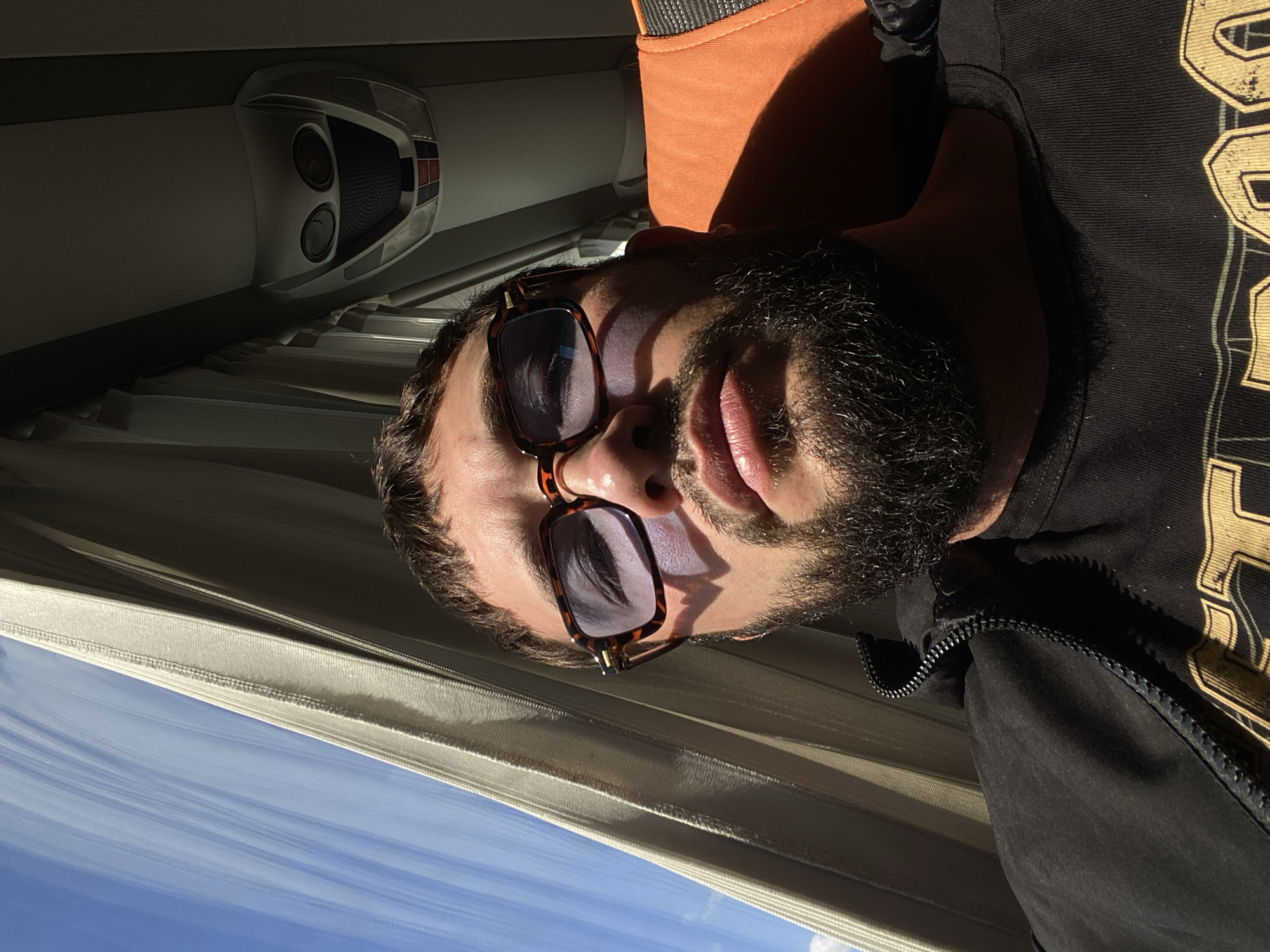 The image shows a close-up, low-angle, selfie-style shot of a man with a beard, wearing sunglasses and a black zip-up jacket over a black t-shirt with a gold graphic design. He is seated on an orange and grey seat, likely in a vehicle such as a bus, as suggested by the window and curtains on the left and the overhead interior on the right. The man's face is illuminated by sunlight streaming through the window. The background shows a blue sky with some clouds and the interior of the vehicle. The location is specified as Glukhovo, Russia.