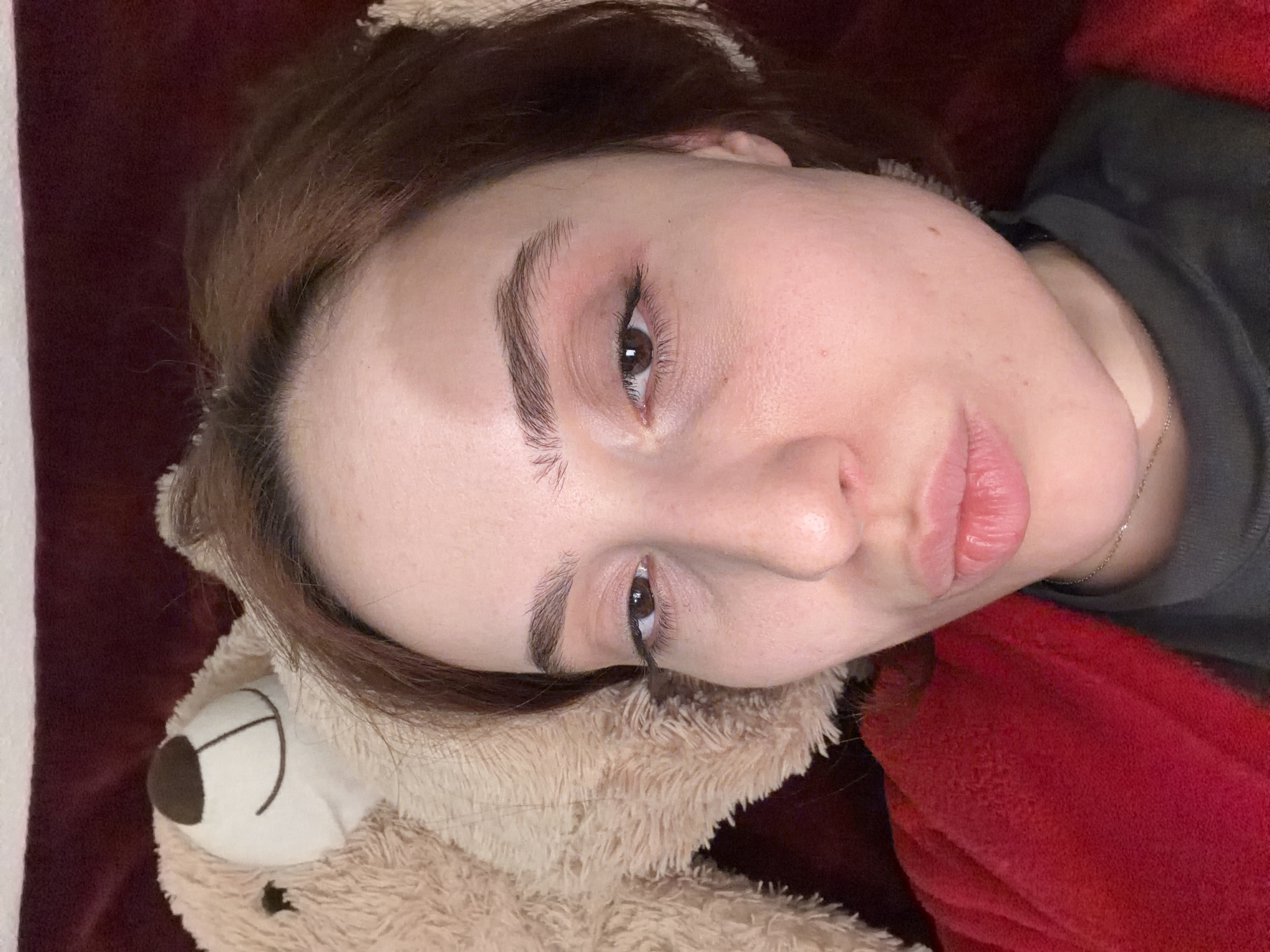 A close-up, eye-level indoor portrait features an adult female subject facing forward. She has light skin, dark brown hair partially visible, and brown eyes exhibiting a slight pinkish shadow. Her lips are pursed in a kissing gesture. She is wearing a dark grey high-neck garment, largely obscured by a textured red fabric, possibly a robe or blanket. A thin, silver-colored chain necklace is visible around her neck. To the subject's left, partially visible behind her head and shoulder, is the light brown, furry head of a teddy bear, displaying a dark brown nose and a stitched smile. The background consists of a dark red or maroon fabric. The image provides no visual indicators to determine its location within the city of Strasbourg, France, or any other specific geographical area.