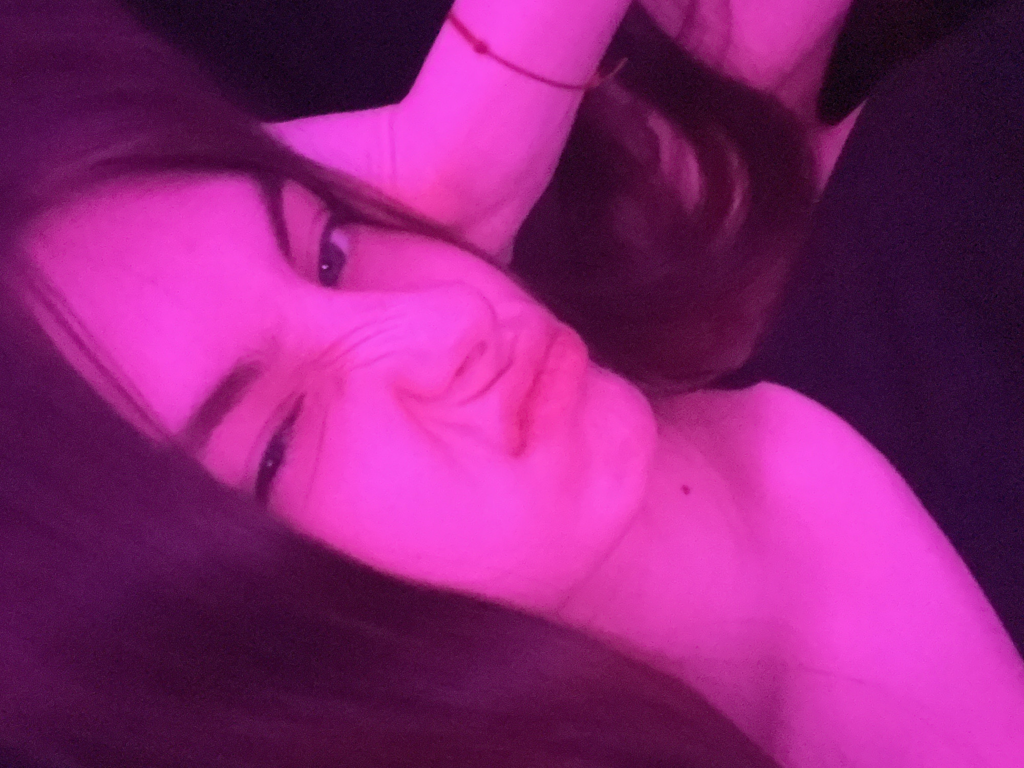 A close-up, angled portrait shows a young woman with fair skin bathed in pink light. She is lying down, her head tilted towards the left, and her right hand is raised near her face, a red string bracelet visible on her wrist. Her facial expression is one of amusement, with scrunched-up features. Long, dark hair frames her face and neck. Part of her shoulder is visible. The background is dark. The image does not provide enough data to determine its location within St. Petersburg, Russia.