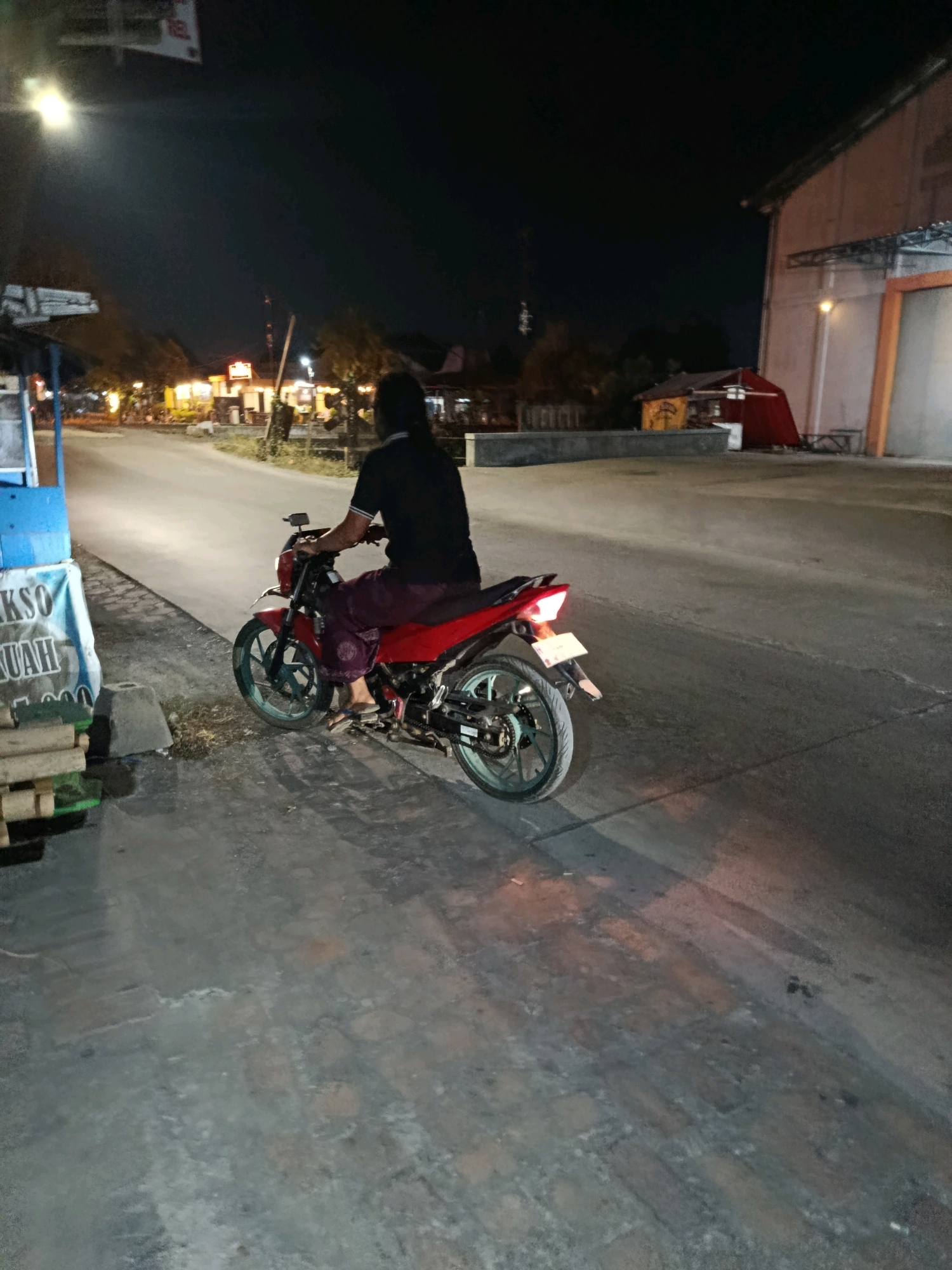 In the city of Tlogorejo, Indonesia, a person is seated on a red motorcycle on a paved road at night. The individual is wearing a dark t-shirt and loose-fitting dark pants, with their back to the camera. The motorcycle has a black seat and a red frame, with light blue rims on the wheels. A license plate is visible on the rear of the motorcycle. The road is illuminated by streetlights and lights from nearby buildings, casting shadows on the ground. To the left of the road, there is a blue and white stall with some wooden crates stacked nearby, and a sign with text that includes "AKSO" and "UAH". In the background, there are buildings and trees, and the sky is dark. The scene appears to be a roadside area with commercial establishments.