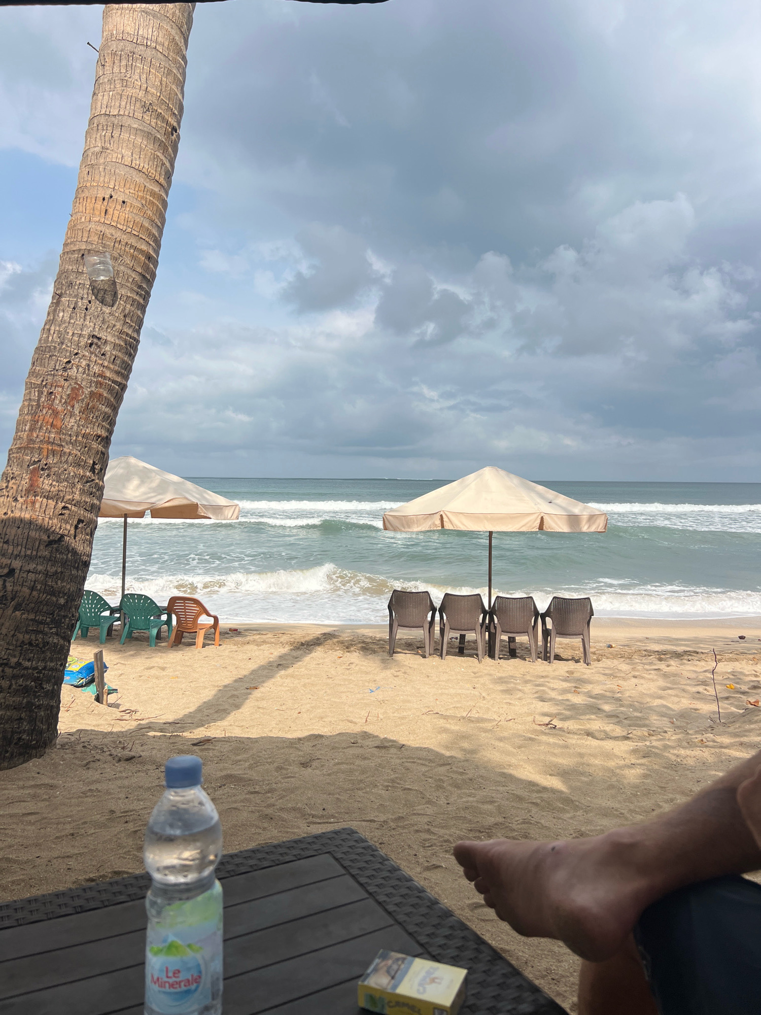 In Kuta, Indonesia, this image captures a beach scene with a prominent palm tree trunk on the left, framing a view of the ocean. The sand is a light tan, with several empty chairs and umbrellas arranged near the water's edge. Two beige umbrellas are set up, one with four dark chairs in front of it and another with three green chairs and one orange chair. The ocean is a light blue-green with moderate waves breaking on the shore under a cloudy sky. In the foreground, a dark table holds a bottle of "Le Minerale" water and a pack of Camel cigarettes. A person's bare foot is visible on the right side of the frame, resting on the table.