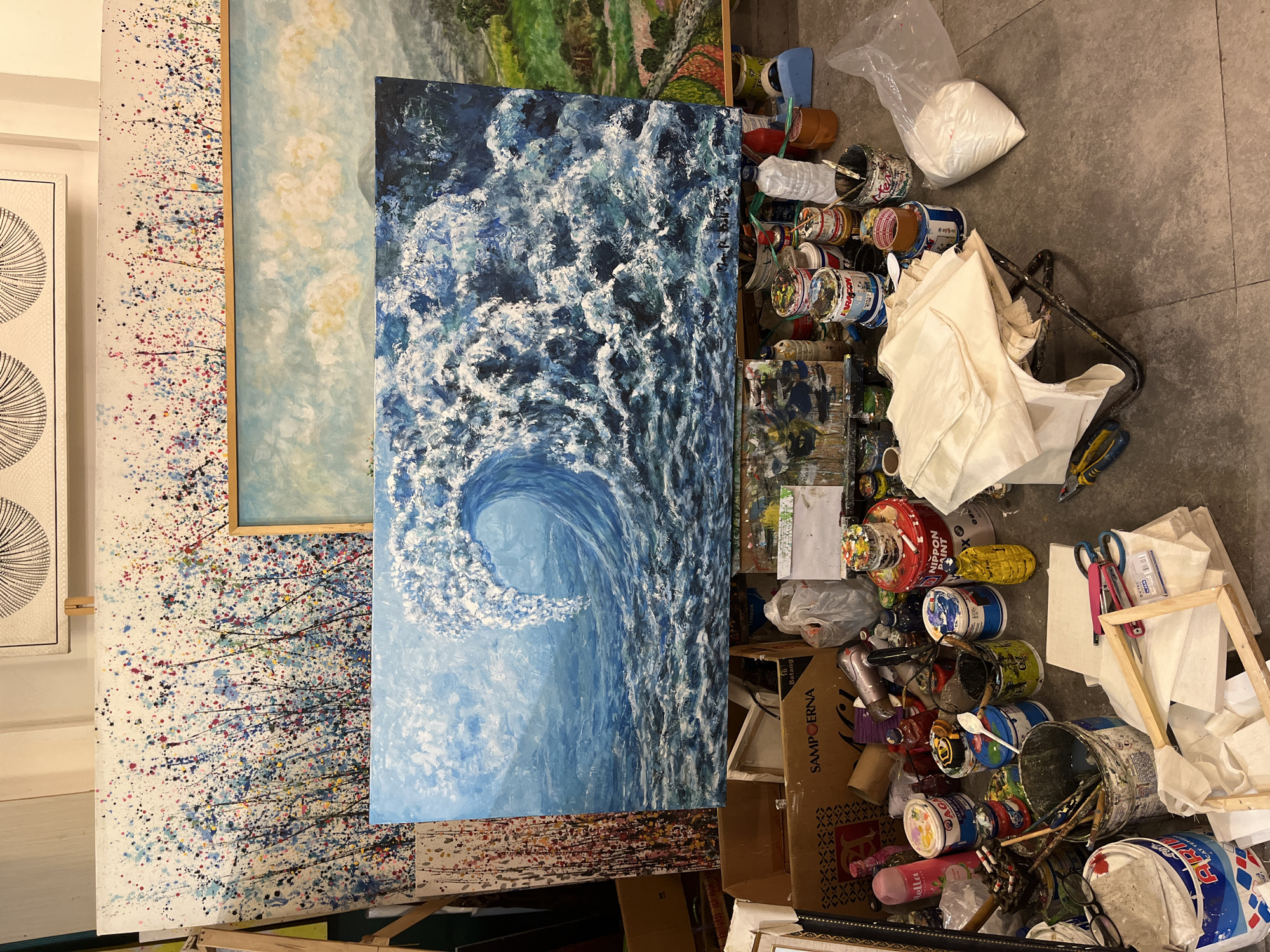 The image shows a collection of paintings and art supplies in a room, possibly a studio. A large painting of a wave dominates the center, with other abstract paintings displayed on easels or leaning against the wall. The floor is cluttered with open paint cans, brushes, tools, and a cardboard box labeled "SAMPOERNA 16 Batang". A white plastic bag and a few cloths are also visible. The room's location is in Kuta Selatan, Indonesia, based on the text "SAMPOERNA 16 Batang," a reference to a cigarette brand produced in Indonesia.
