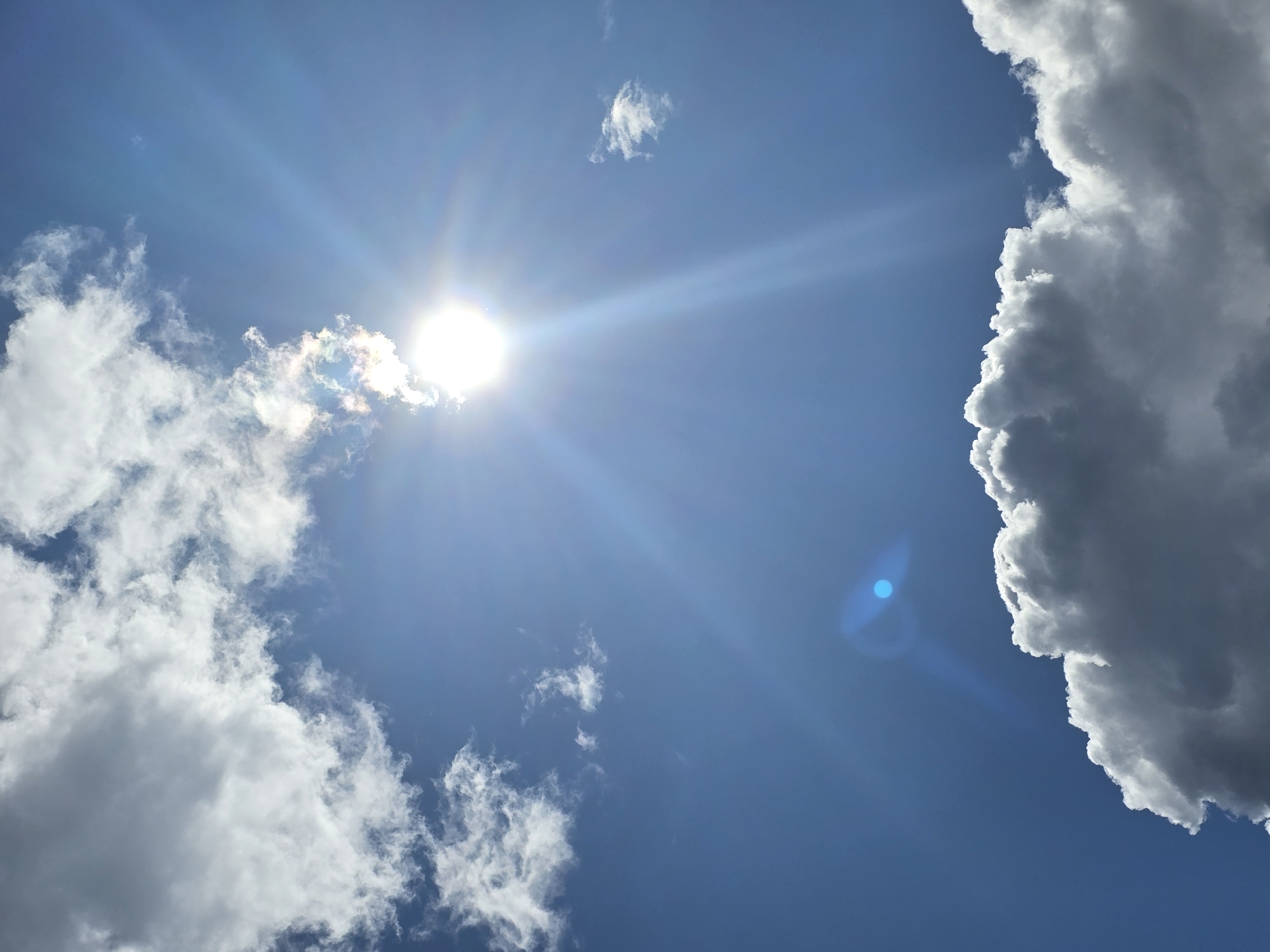 A bright sun shines through a clear blue sky in Ермолаево, Russia, casting rays of light. White, fluffy clouds are scattered across the sky, with some larger formations near the bottom of the frame. One cloud near the sun appears to have a rainbow halo around it. A blue lens flare is visible in the lower left quadrant of the image. The scene depicts a sunny day with some cloud cover.