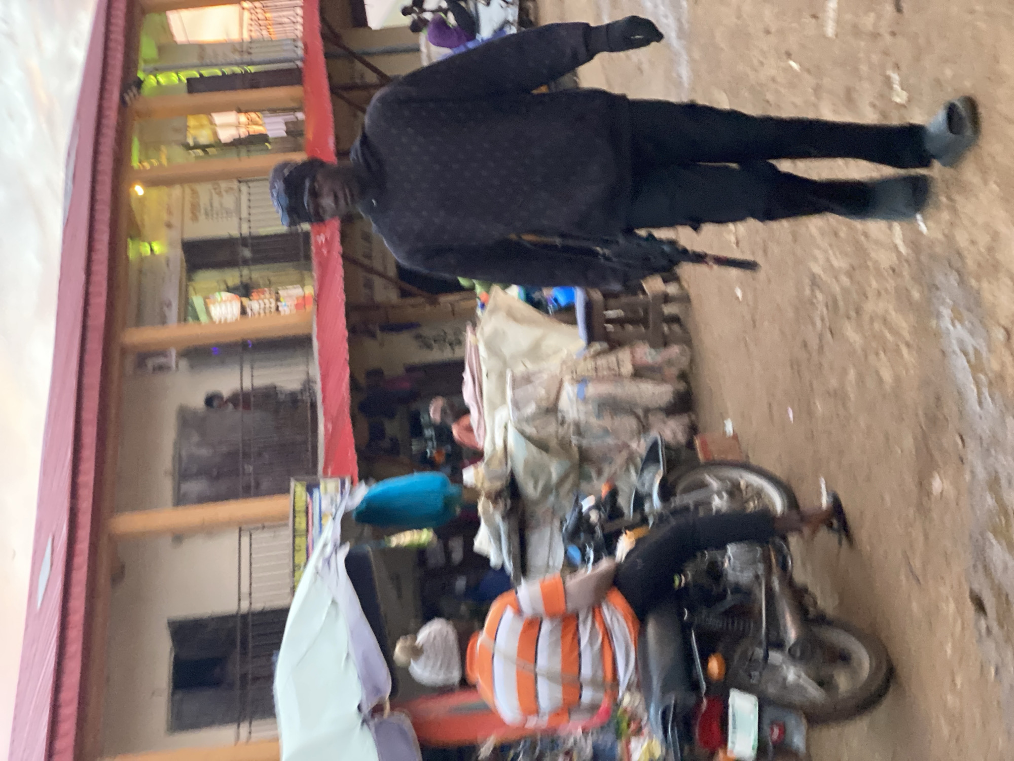 The image depicts a street scene in Kafanchan, Nigeria, with two individuals prominently featured. In the foreground on the right, a person walks towards the camera, wearing a dark hooded sweatshirt with a patterned design, dark pants, black gloves, and dark footwear. They are carrying a dark object that resembles a rifle in their right hand. On the left side of the image, another person is seated on a motorcycle, facing away from the camera. This individual is wearing a horizontally striped orange and white t-shirt and black pants, with a white knitted hat. The motorcycle is parked on a dirt or gravel surface. Behind the individuals, there is a building with a red roof, exhibiting multiple stories with balconies and open doorways. The ground appears to be wet and muddy in places, suggesting recent rain. The overall environment suggests an outdoor market or commercial area.