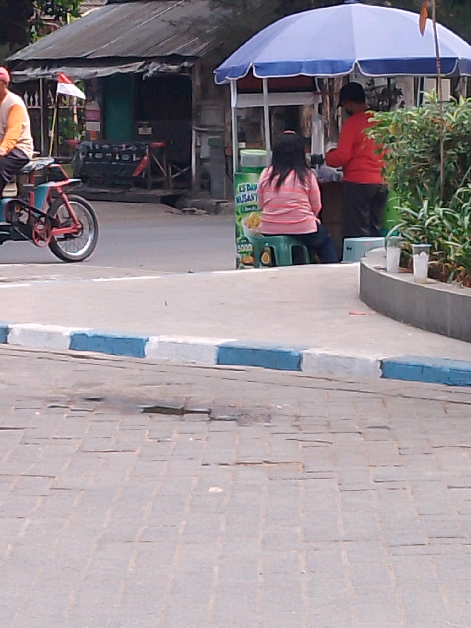 In Kutorejo, Indonesia, an outdoor scene depicts a food stall and its surroundings. A man, wearing a red long-sleeved shirt and dark pants, stands behind the counter of a blue and white umbrella-covered food stall, appearing to serve a customer. A woman, seated on a green stool facing away from the camera, wears a pink and white striped shirt and dark pants, and is looking towards the stall. A red bicycle with a blue cargo carrier is parked to the left of the scene. In the background, a small, weathered building with a dark roof and a white and red flag is visible. The ground is paved with grey cobblestones.