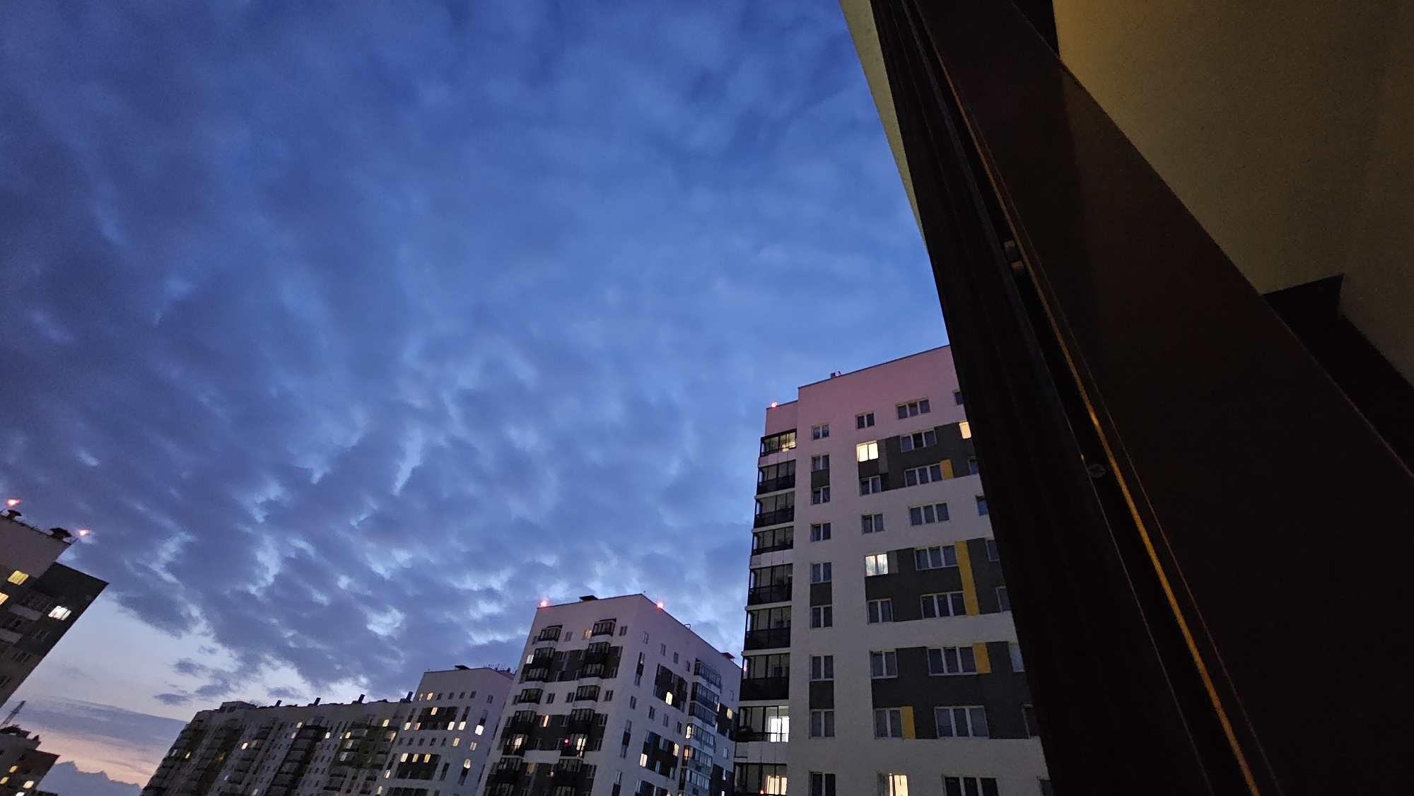 The image captures a twilight view of several apartment buildings in the city of Yekaterinburg, Russia. The sky is a deep blue with scattered clouds, illuminated by the fading light of dusk. Several buildings are visible, with windows lit from within, suggesting it is evening or nighttime. The architecture of the buildings appears modern, with multiple stories and balconies. A dark, vertical structure, possibly a part of a building or a balcony railing, occupies the right side of the frame, obscuring a portion of the scene. The lighting suggests artificial light sources within the apartments are the primary illumination for the buildings. No people or specific actions are depicted. The scene is static and conveys a sense of urban residential life at the end of the day.