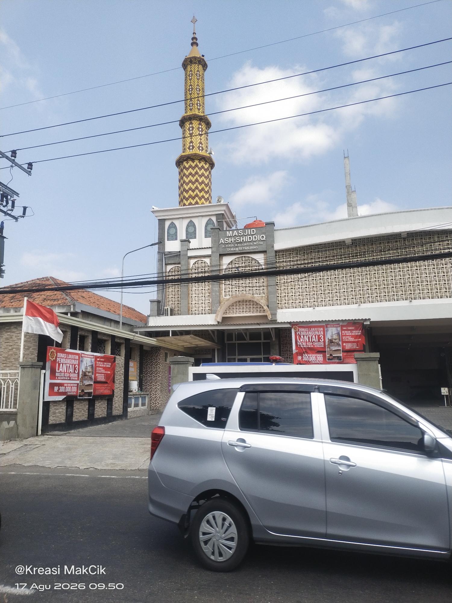 In the city of Kalijaten, Indonesia, on August 17, 2026, at 09:50, a silver minivan is parked on the street in front of the Masjid Ash-Shiddiq. The mosque features a tall, golden minaret with brown patterned accents, and the building itself has a modern design with decorative window elements. To the left of the mosque, a red and white Indonesian flag flies above a building with a tiled roof. Two red banners are displayed on the left side of the mosque, one of which advertises "DONASI LANJUTAN PEMBANGUNAN LANTAI 3 MASJID ASH-SHIDDIQ" (Donation for the continuation of the 3rd floor construction of Ash-Shiddiq Mosque), with a target amount of RP. 300.000.000. Overhead, power lines are visible against a clear blue sky with scattered clouds. The image is watermarked with "@Kreasi MakCik".