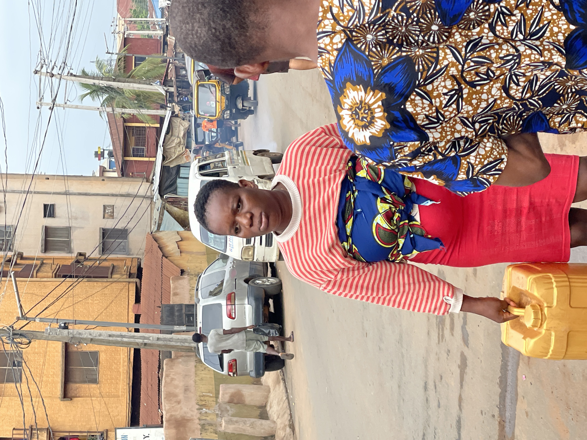 A woman in a red and white striped shirt and red skirt carries a yellow jerrycan in a street in Onitsha, Nigeria. A blue and brown floral patterned cloth is draped over her shoulders. She walks towards the viewer, facing forward. Another person in a matching blue and brown floral patterned shirt is partially visible in the upper right corner. A white van, a silver SUV, and a yellow motorized tricycle (keke) are visible in the background on the street. Buildings are on either side of the street, with visible power lines overhead. A man walks along the side of the street near the silver SUV.