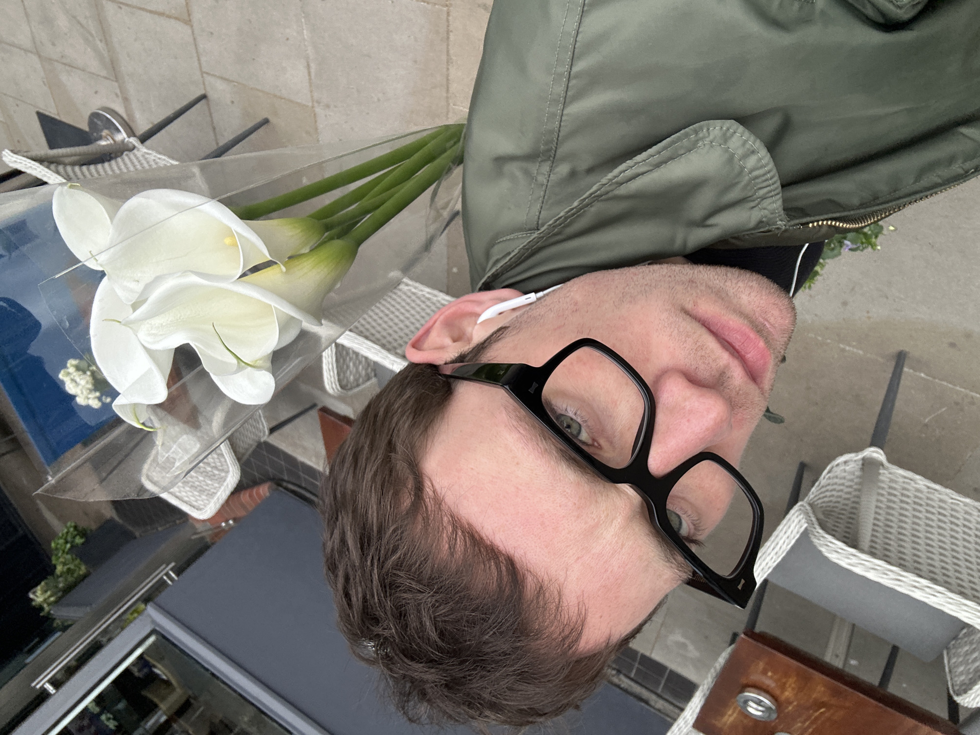 The image shows a man wearing black-rimmed glasses and a green jacket, holding a bouquet of white calla lilies wrapped in clear plastic. He appears to be seated outdoors, possibly at a cafe or restaurant in London, United Kingdom. The background includes a woven chair, a table, and glimpses of the city environment, suggesting an urban setting.