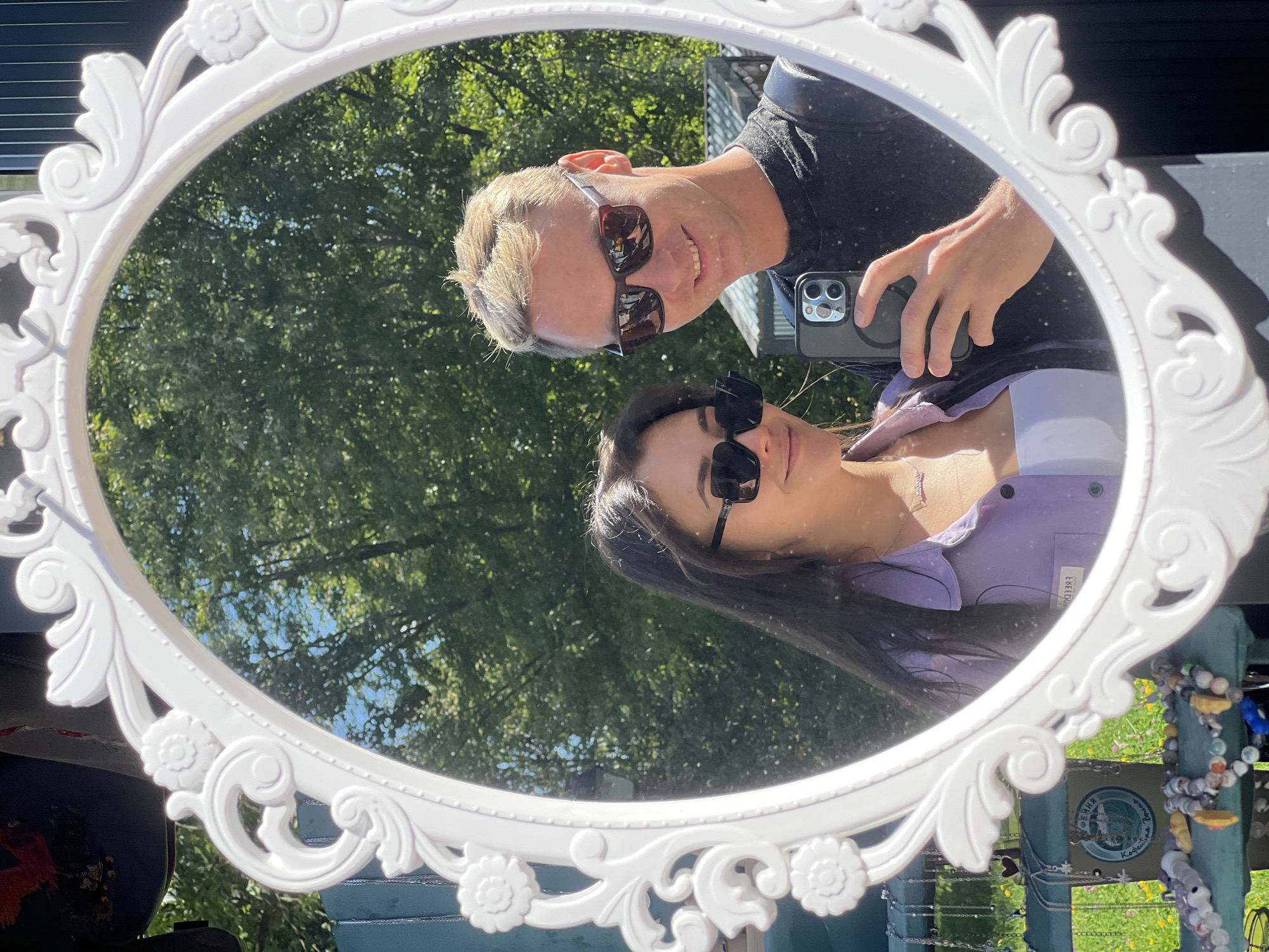 In the reflection of an ornate white oval mirror, a young Caucasian man and woman are taking a selfie outdoors on a sunny day. The man, on the right, has blonde hair, sunglasses, and is wearing a black t-shirt. He is holding a smartphone, likely capturing the image. The woman, on the left, has long dark hair, sunglasses, and is wearing a light purple open button-down shirt over a white top. She is smiling. Behind them, lush green trees form a dense backdrop, suggesting a park or outdoor market setting. The mirror's frame is decorated with floral and scroll motifs. In the foreground, visible to the left and below the mirror, are items displayed on a blue surface, possibly from a market stall, including necklaces and other decorative objects. The overall ambiance is casual and bright, indicative of a pleasant outdoor outing.