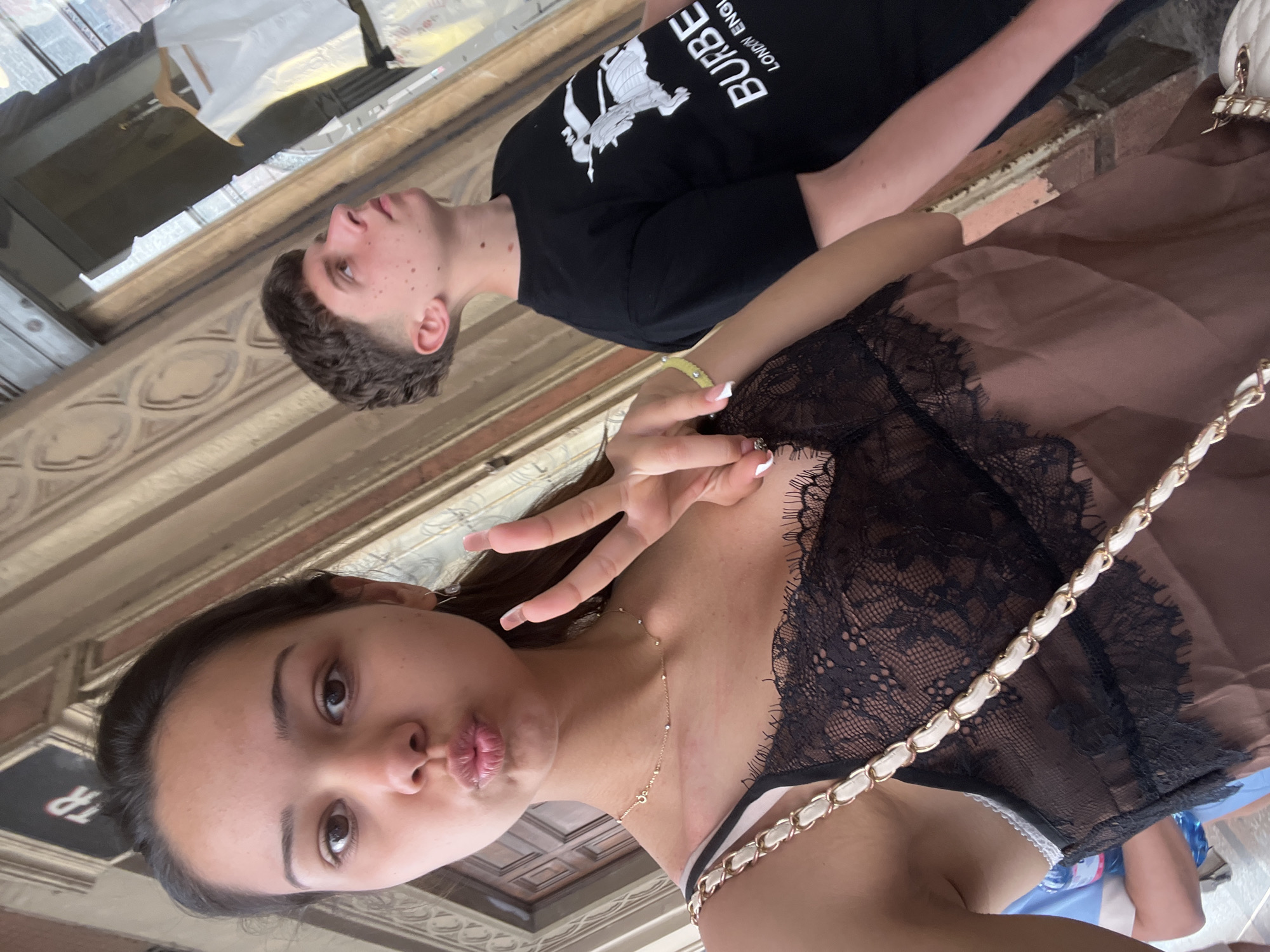 The image appears to be a selfie taken indoors. A young woman is in the foreground, making a kissing face and a peace sign with her hand. She has brown hair and is wearing a black lace top with a gold necklace. A purse strap with a white and gold chain is visible across her chest. Behind her is a young man wearing a black t-shirt with white lettering. The background shows architectural details suggesting an older building, possibly a museum or historical site, with stone walls and patterned ceilings.