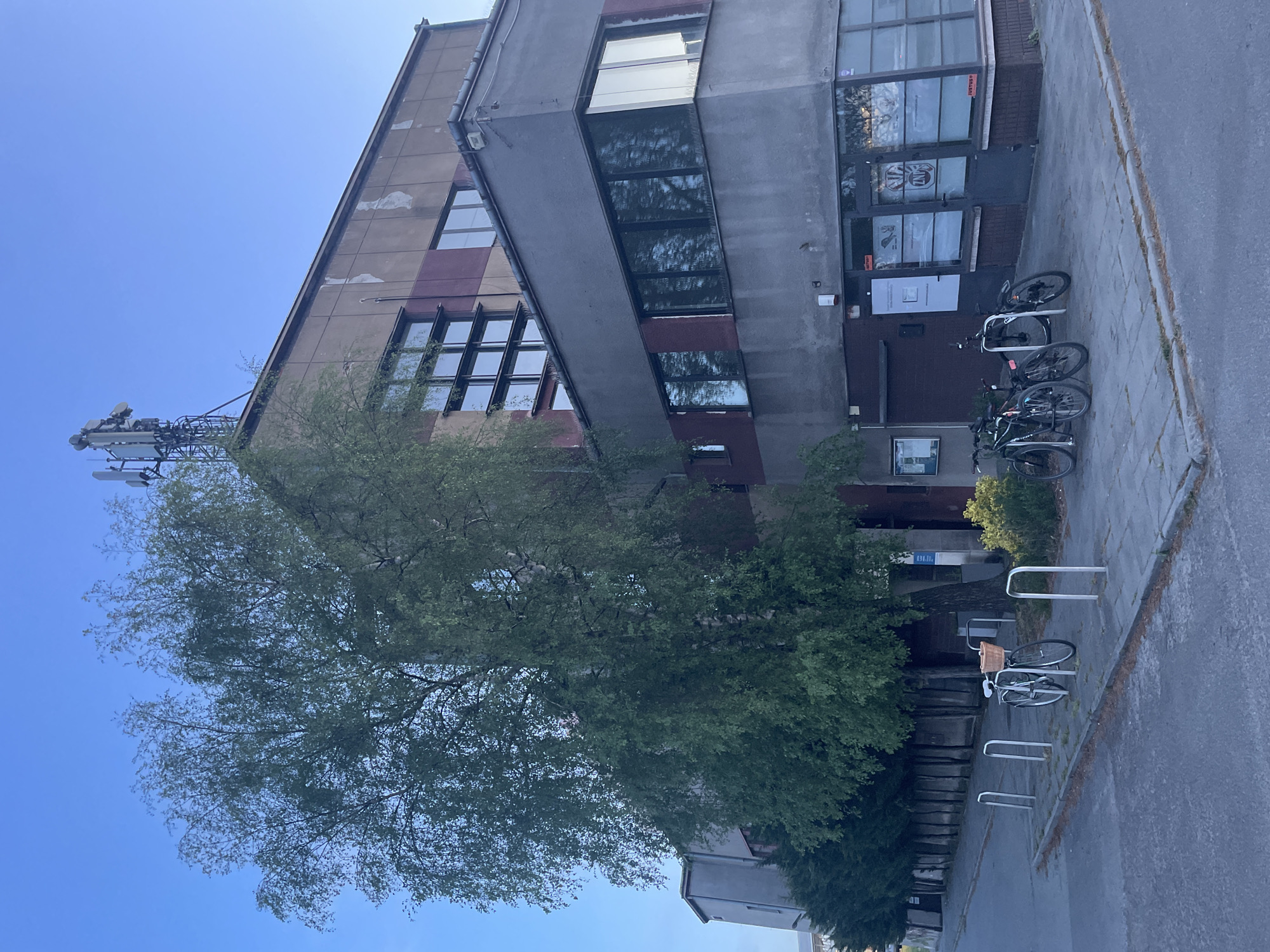 The image depicts a multi-story building with a modern, angular design. The building has a combination of concrete and windowed sections. A large tree is located in front of the building, partially obscuring the view of the lower floors. A telecommunications tower is visible on the roof of the building. Several bicycles are parked in front of the building, near a sidewalk. The location appears to be an urban area with clear skies.
