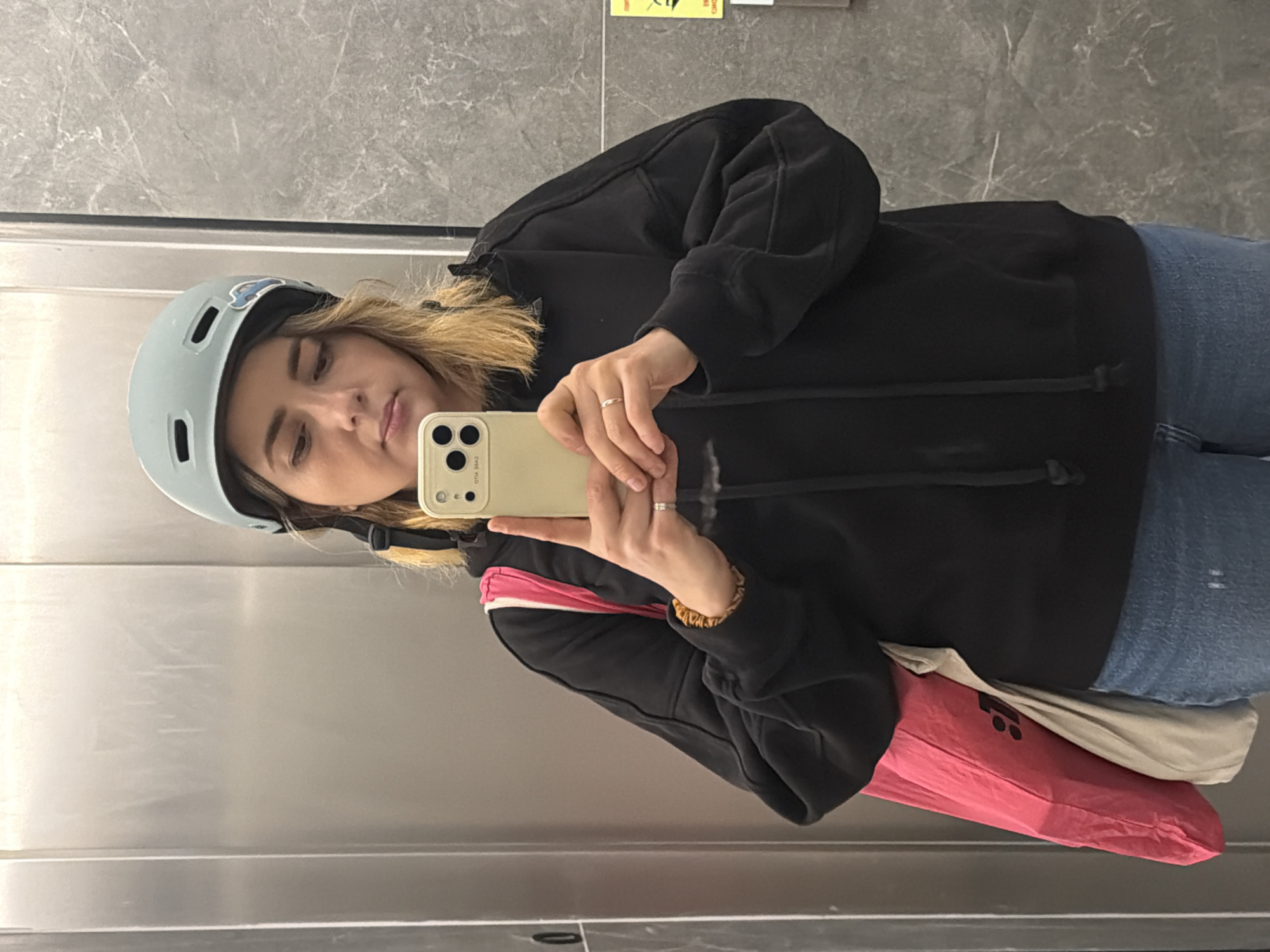 A young woman is taking a selfie in what appears to be an elevator. She is wearing a light blue helmet, a black hoodie, and blue jeans. She is holding a beige-colored smartphone, likely an iPhone, in her right hand, and her reflection is visible in the stainless steel or polished metal walls of the elevator. A pink bag is visible slung across her body. The background is the interior of the elevator, featuring gray marble-like wall panels and a yellow warning sign. Without further information, it's impossible to determine the precise location in Ramat Gan, Israel.