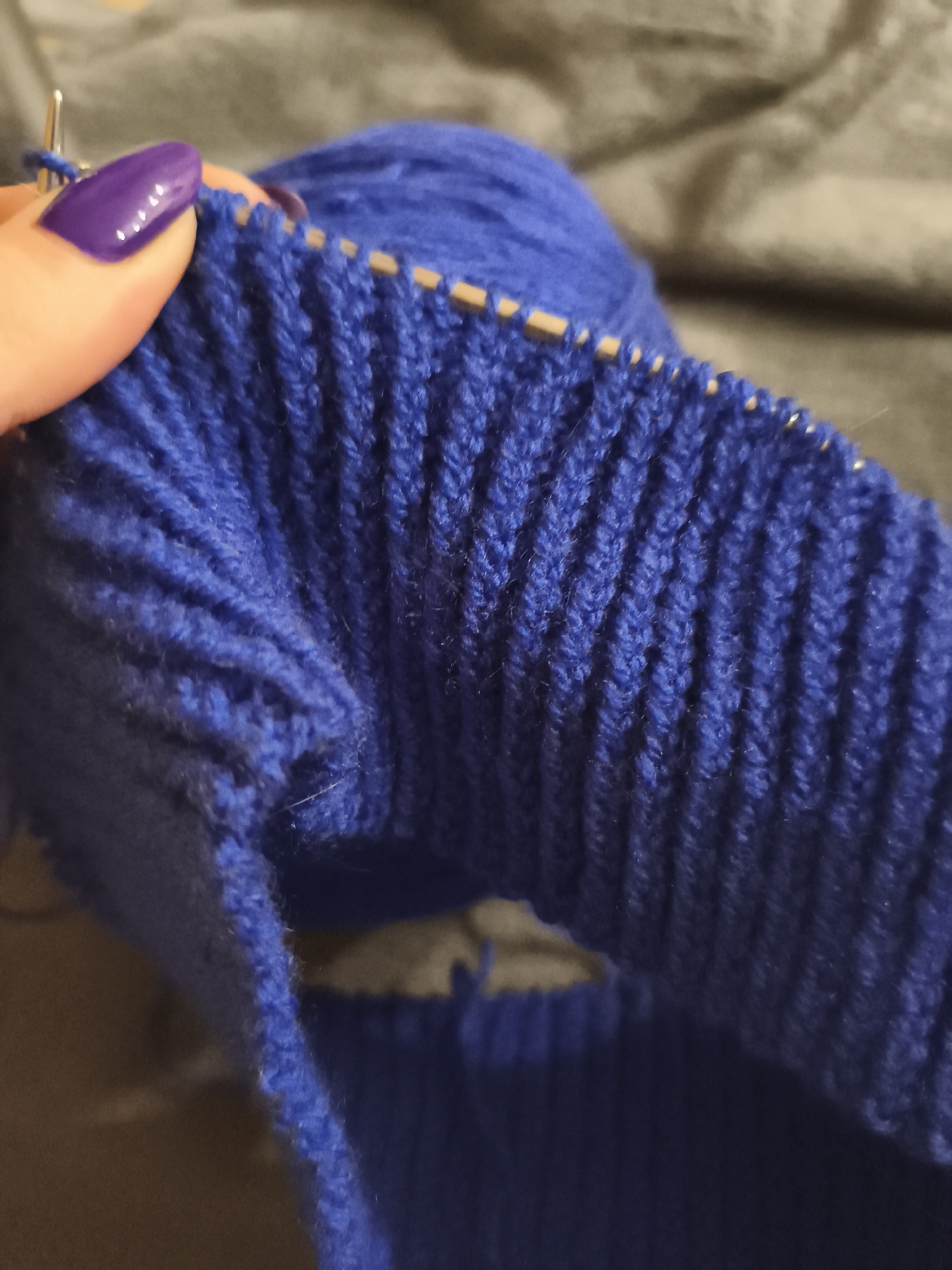 A close-up shot shows a person knitting a blue garment. The garment appears to be ribbed, and the knitting needles are visible with stitches on them. The person's hand, with purple nail polish, is holding the knitting. A ball of blue yarn is in the background, resting on a grey surface. There is no indication of the location being Русский Вожой, Russia.