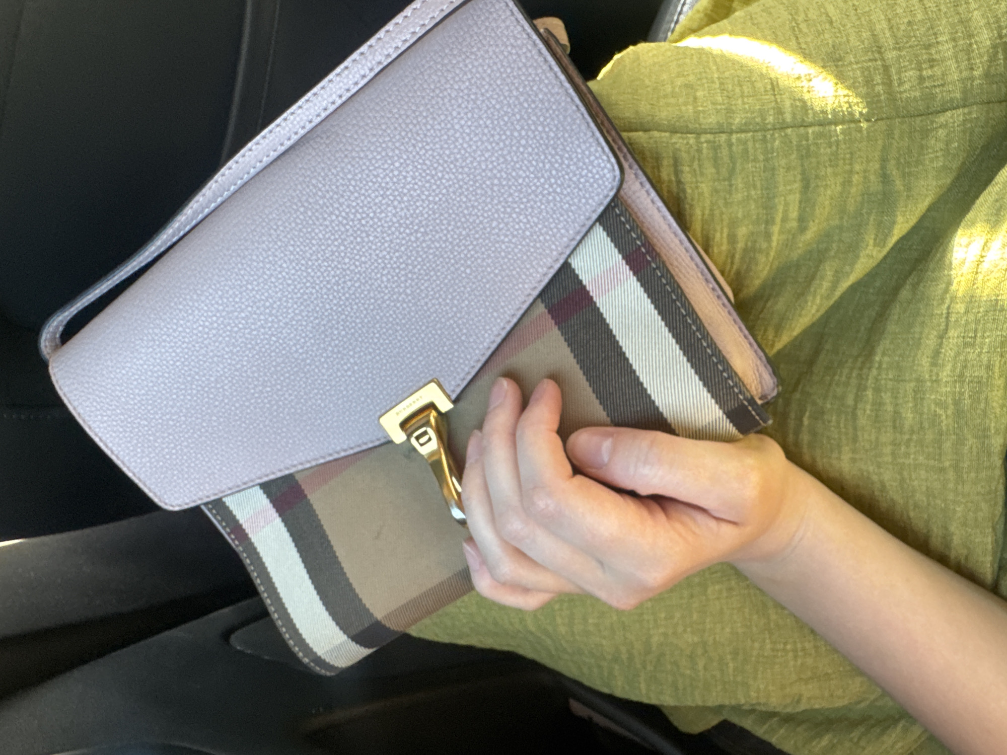 A person's left arm and hand are visible, holding a light purple leather clutch purse with a gold clasp. The clutch is resting on top of a beige and brown checkered fabric that appears to be another bag or part of the person's clothing. The person is wearing a textured, lime green garment, possibly a dress or skirt. The background is dark and indistinct, suggesting an indoor or shaded environment. The lighting indicates it is daytime.