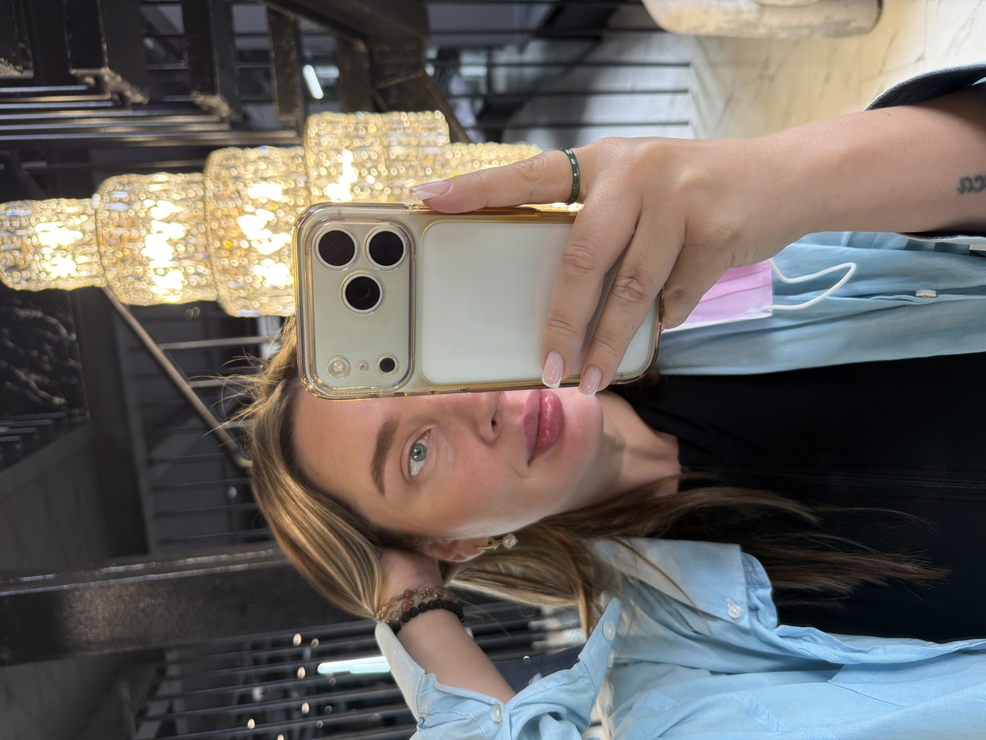 A woman is taking a selfie in front of a chandelier. The woman has light hair with highlights and light eyes. She is wearing a light blue shirt and a black top. She is holding a gold iPhone with a clear case. The chandelier is large and has multiple tiers of lights. The background is blurry, but it appears to be a building. The image was taken in Moscow, Russia.