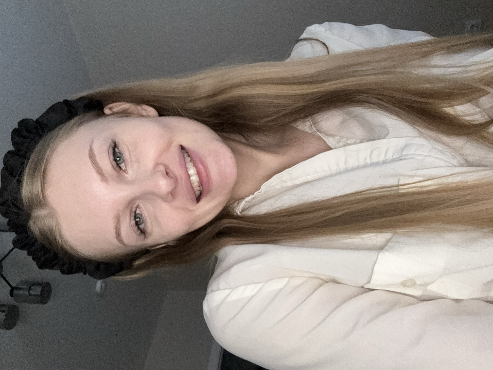 A young woman with long blonde hair and a black headband smiles at the camera. She is wearing a white collared shirt. The background is a plain gray wall. The image appears to be taken indoors, possibly in a residential setting in the city of Perm, Russia.