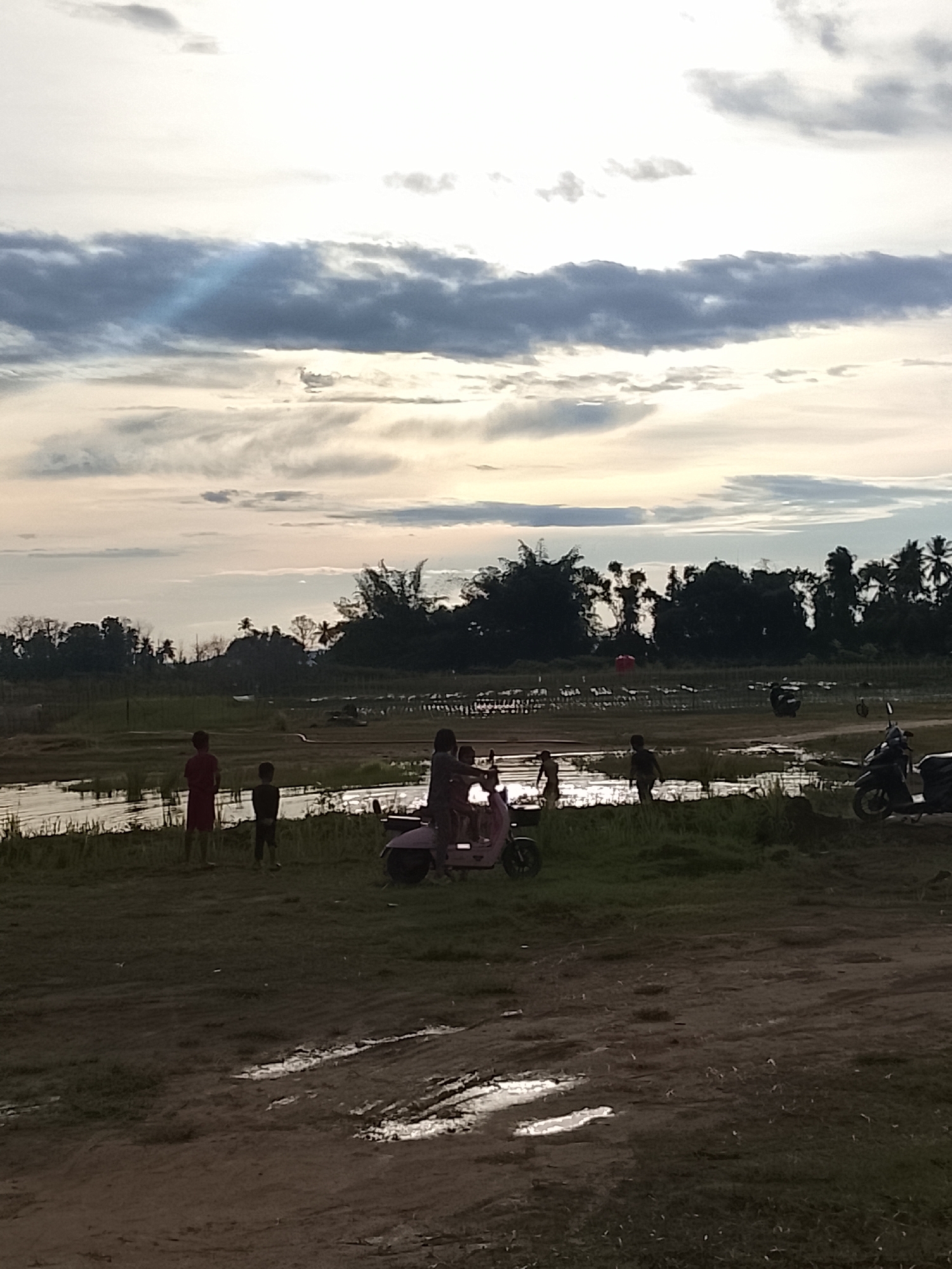 A wide shot captures a muddy, outdoor scene in Geunteng, Indonesia, under a cloudy sky. In the foreground, a dirt path with several puddles indicates recent rain. The middle ground features a grassy area bordering a shallow body of water or flooded field. Several children are present in this area. Two boys stand near the water's edge, one wearing a red shirt and the other a dark shirt. Further back, a light-colored scooter is occupied by at least two children, one seemingly driving. To the right, a dark-colored scooter is parked. In the background, a dense line of trees, including palm trees, forms a dark silhouette against the lighter sky. The sky itself is filled with a mix of dark and light clouds, with a faint ray of light breaking through the clouds in the upper left quadrant.