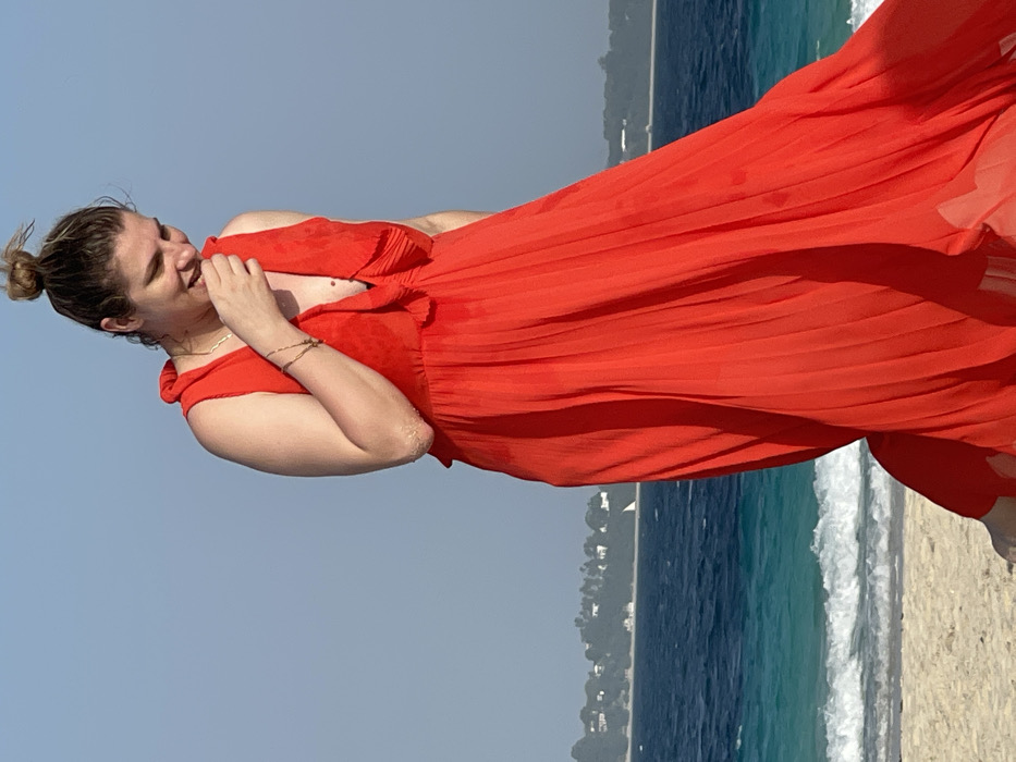 An adult female is depicted standing on a sandy beach along the coast of Zanzibar City, Zanzibar, Tanzania. She wears a sleeveless, bright red pleated maxi dress with a ruffled V-neckline. Her hair is gathered into a bun on top of her head. Her head is tilted downwards, and her right hand is positioned near her mouth, indicating an expression of amusement or shyness. Multiple delicate bracelets are visible on her left wrist, and sand residue is present on her left elbow. In the background, the Indian Ocean displays varying shades of blue and turquoise, with white-capped waves breaking on the shore. A distant green coastline dotted with white structures is visible across the water under a clear blue sky.