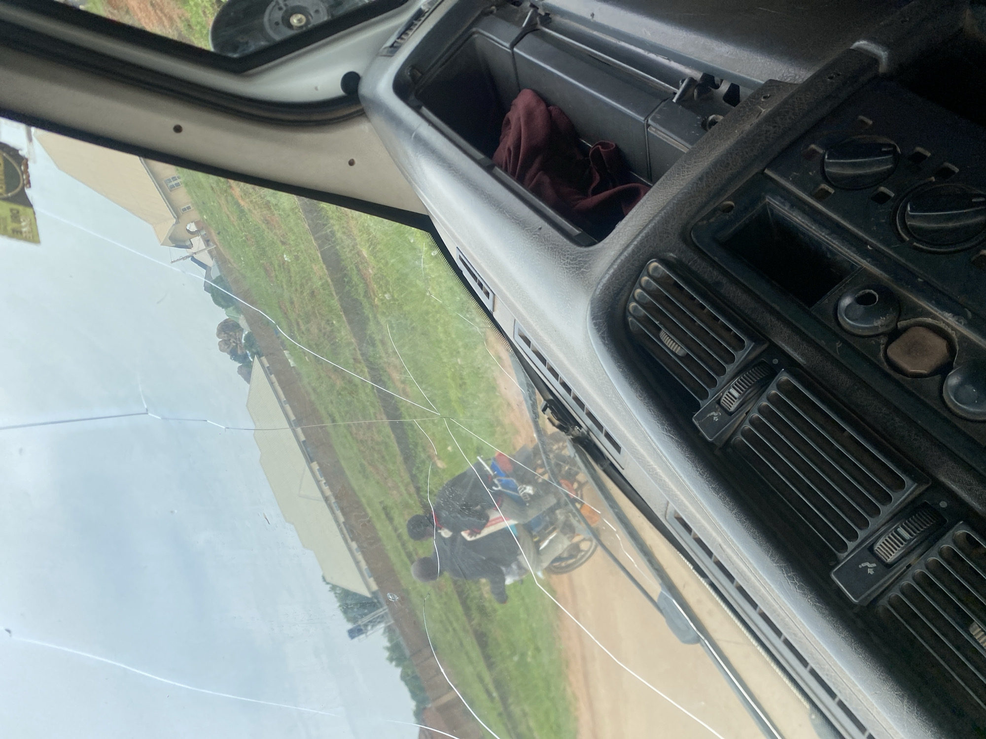 The image was taken from inside a vehicle, looking through a cracked windshield. The view outside shows a dirt road with a house on a grassy hill in the background. Two people are riding on a motorcycle on the road, heading away from the camera. The interior of the vehicle is visible in the foreground, including the dashboard with air vents and controls. The overall scene suggests a rural or semi-urban environment in DU, Nigeria, possibly on a cloudy day.