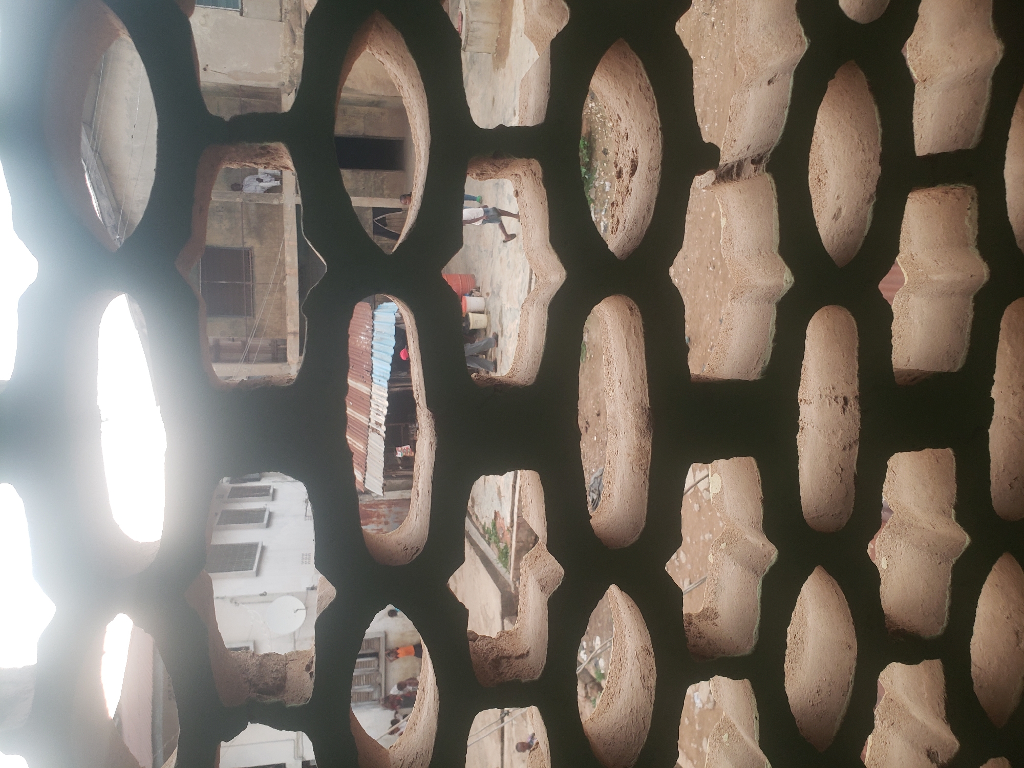 The image depicts a perforated wall, likely made of a porous, earthen material, providing a blurred and fragmented view of the surrounding urban environment in Alalubosa, Nigeria. The wall is composed of regularly spaced, oval and hourglass-shaped openings. Through these openings, buildings with various architectural styles are visible, including structures with corrugated metal roofs and white facades with windows and satellite dishes. People can be discerned in the background, engaged in everyday activities. The overall impression is one of a densely populated, lived-in urban area, viewed through the obscuring filter of the perforated wall. A strong light source is present, causing glare in some openings.