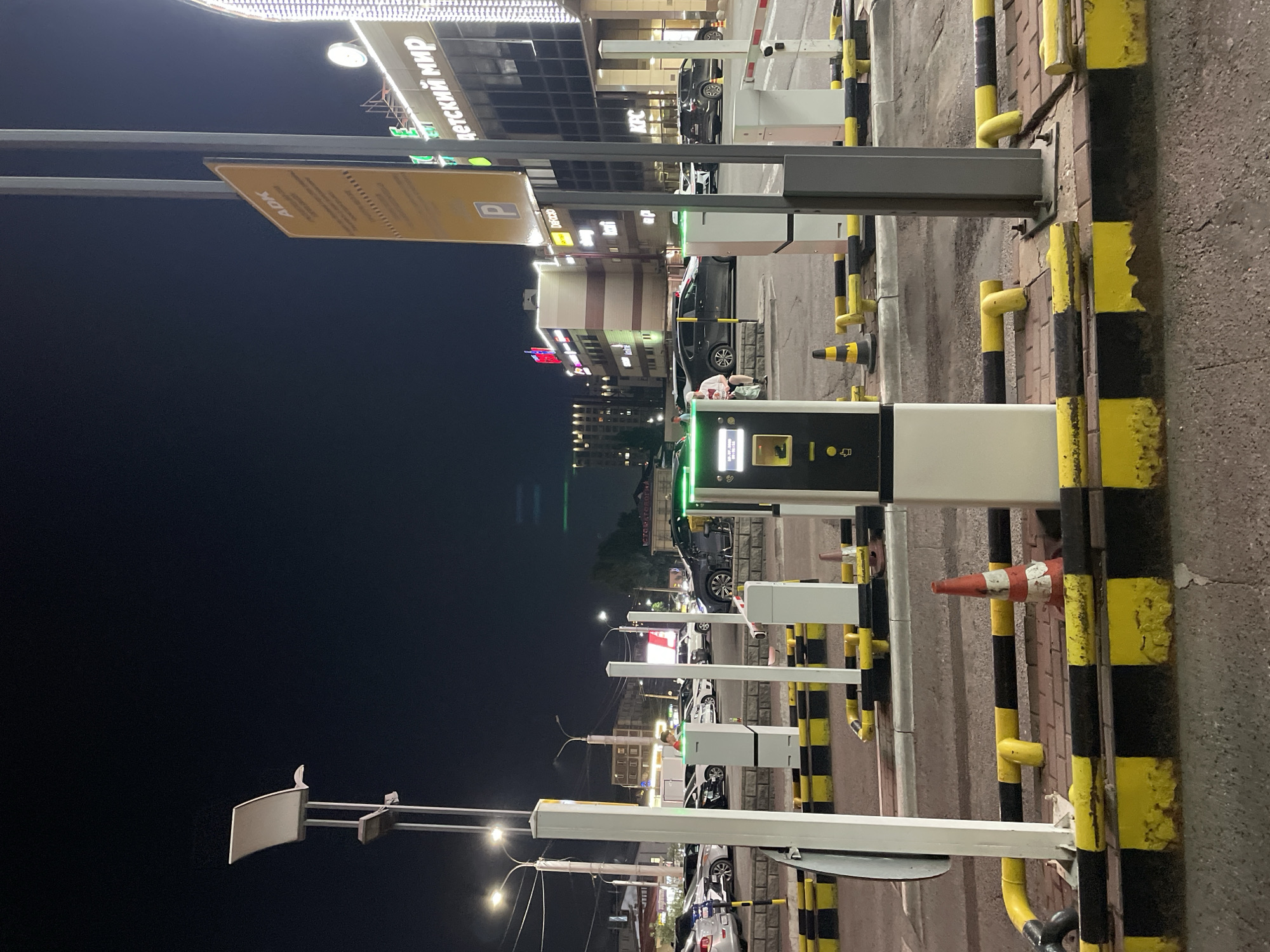 A nighttime exterior shot captures a parking lot in Almaty, Kazakhstan, with a focus on automated payment terminals and barriers. The scene is illuminated by artificial lights from streetlamps and the facade of a large building to the right, which displays illuminated signs, including "ДЕТСКИЙ МИР" (Children's World). In the foreground, a white parking payment terminal with a touch screen and ticket dispenser is prominent. Adjacent to it are white metal posts and yellow and black striped barriers indicating parking spaces. Several cars are parked in the background, and a few people are visible near the payment terminals. The ground is paved with asphalt and brick. A traffic cone with orange and white stripes is positioned near a barrier.