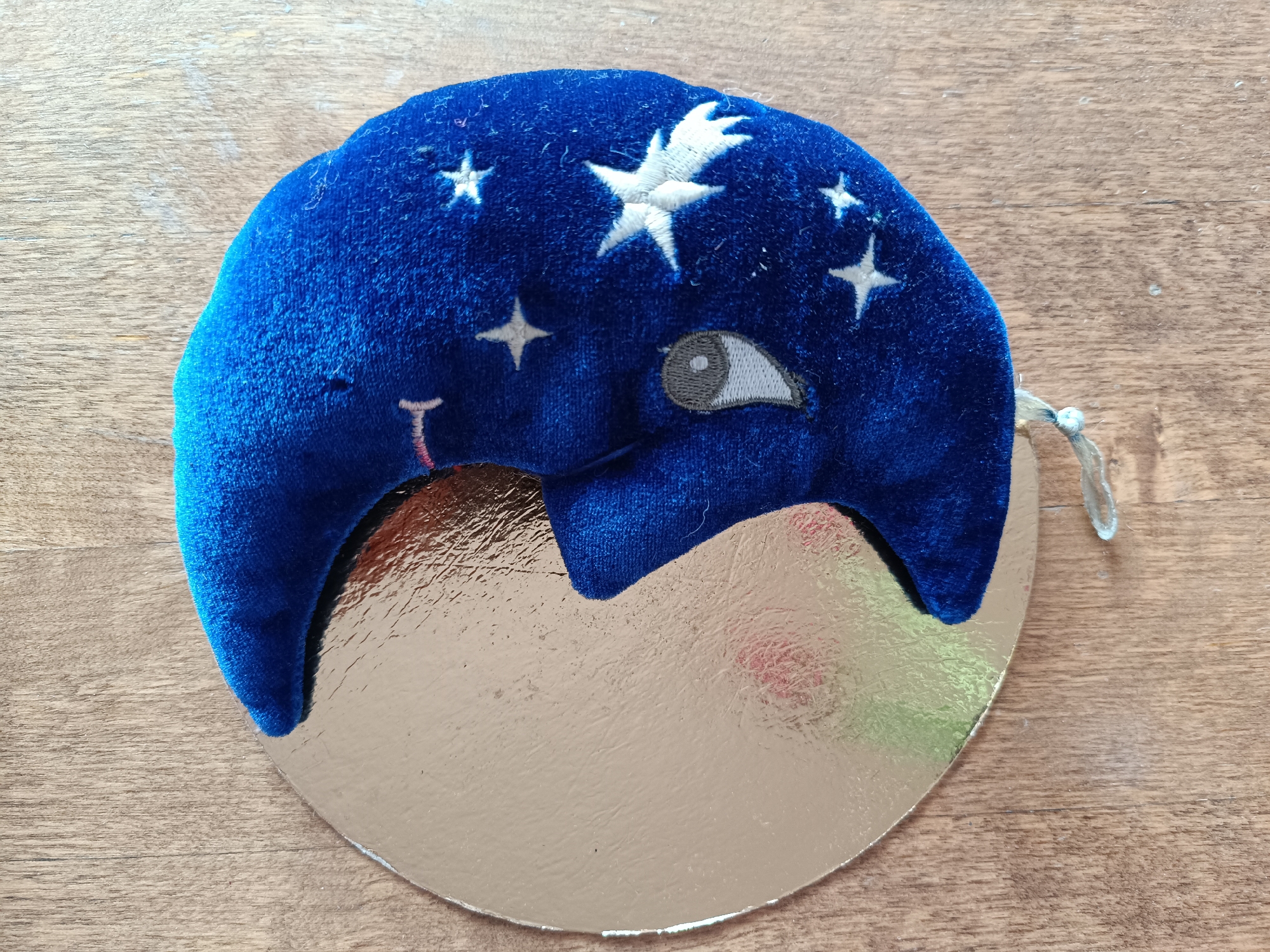 A dark blue velvet, crescent moon-shaped object rests on a circular, reflective metallic surface, which in turn is placed on a light brown wooden background. The velvet object is adorned with several embroidered white stars of various sizes across its upper section, a stylized embroidered eye with a gray iris and white sclera on its right side, and a small pink, inverted T-shaped embroidery on its left. A thin, twisted white cord loop is attached to the upper right edge of the velvet object. The reflective surface beneath the velvet, likely a metallic cake board, exhibits minor surface undulations and reflects ambient light, creating subtle color shifts. The wooden background displays a visible grain pattern, light greyish-brown coloration, and minor textural inconsistencies. This scene is situated in the city of Tebras, Latvia.