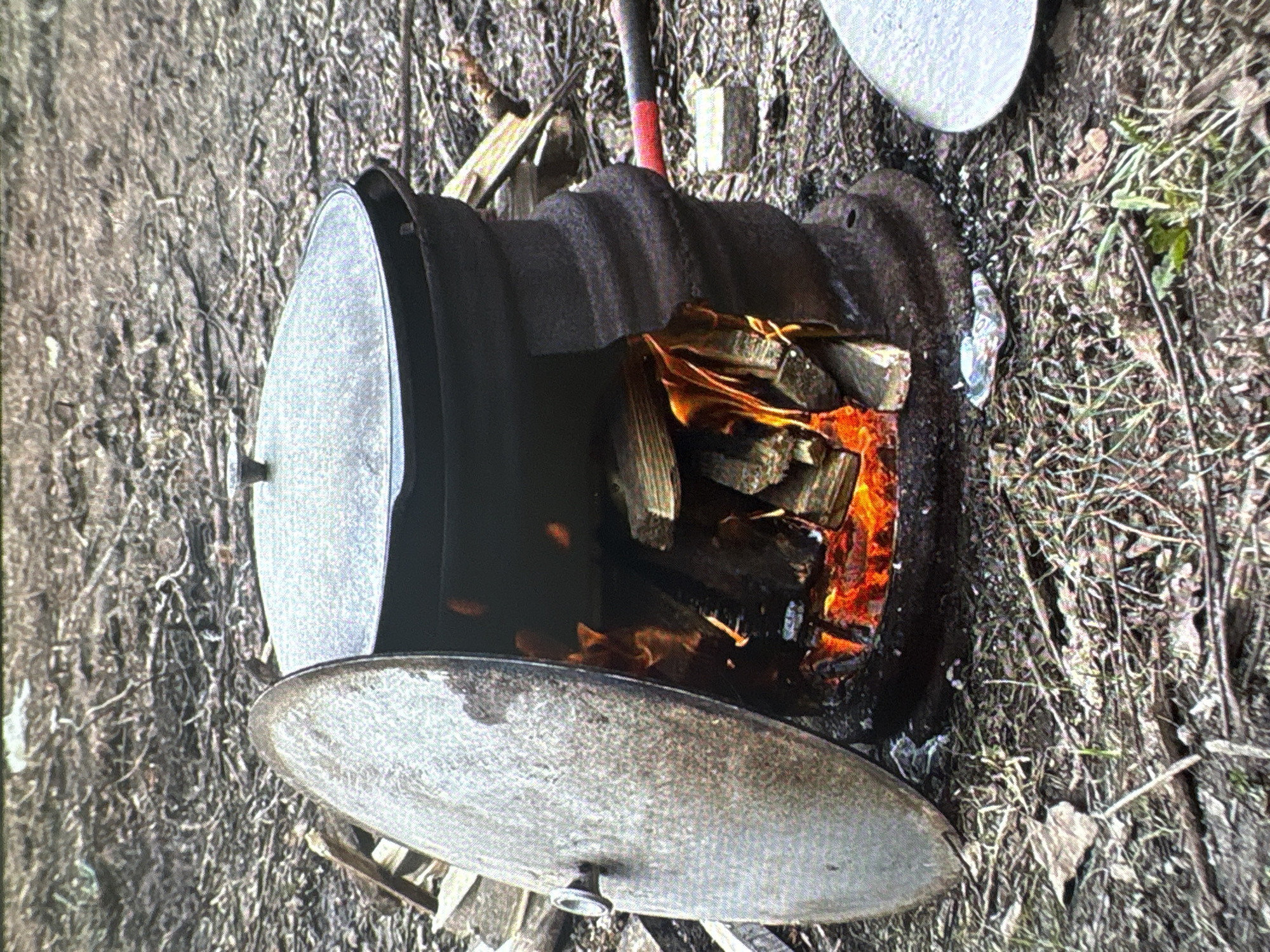 An outdoor scene in Batumi, Georgia, shows a rustic cooking setup. A large, dark cauldron with a lid ajar sits over an open fire, fueled by logs. Flames are visible within the cauldron's opening. A large metal lid is propped open to the left of the cauldron. The ground is covered with dirt, dry leaves, and twigs. To the right of the cauldron, a portion of another metal lid is visible. An object with a red handle and a black head, resembling a shovel or a tool, is placed behind the cauldron. The overall impression is of a makeshift cooking area in a natural or semi-natural outdoor environment.
