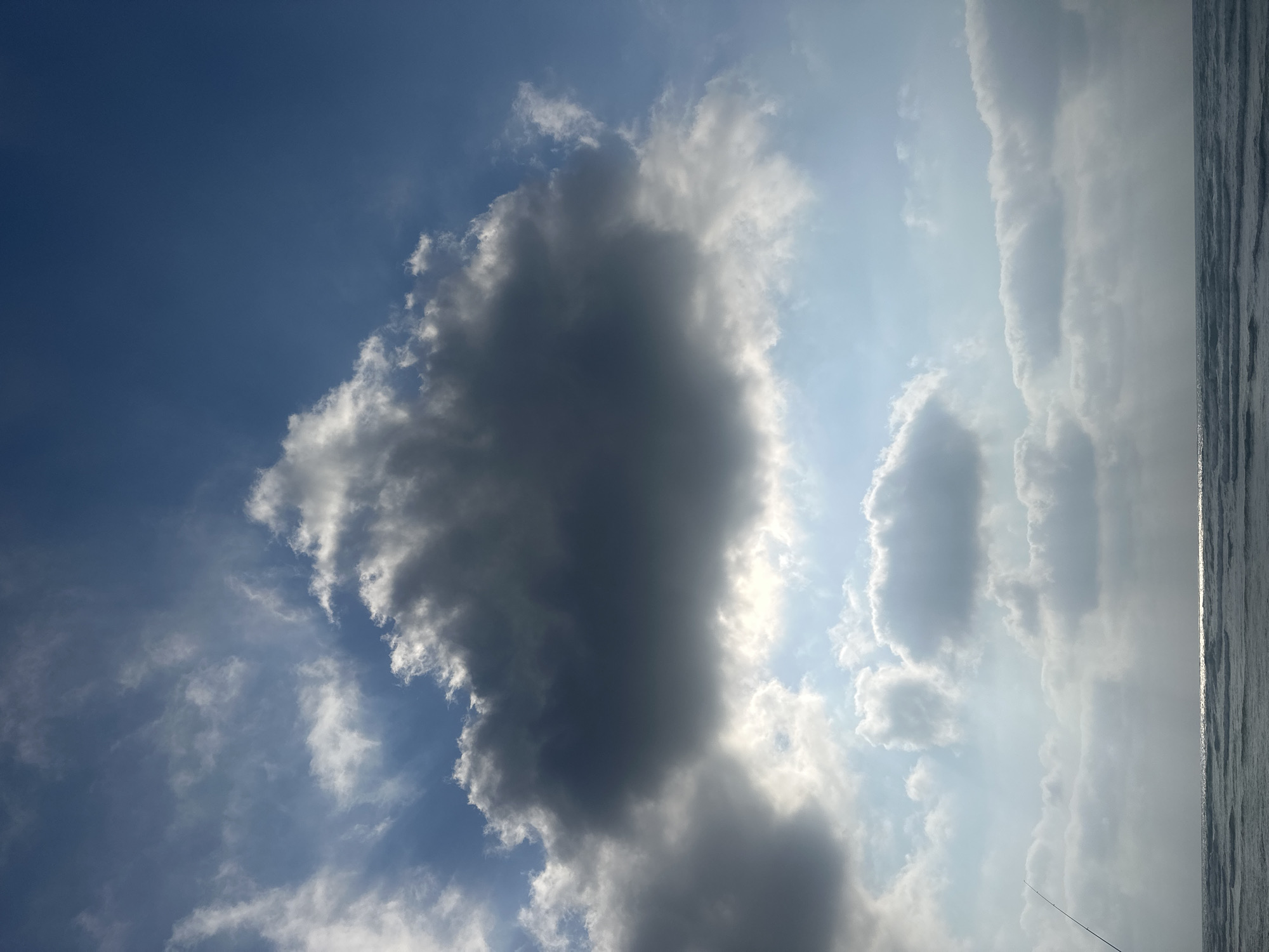 The image captures a view of the Mediterranean Sea from Haifa, Israel. A fishing rod is visible in the lower left corner, extending upwards at an angle, suggesting a person is fishing. The sea is relatively calm with gentle waves and a glint of sunlight on the water. Above the sea, the sky is a mixture of blue and dramatic clouds. A large, dark grey cloud dominates the upper center of the frame, while lighter, white clouds are scattered across the sky, some illuminated by sunlight breaking through. The overall scene depicts a tranquil coastal environment under a partly cloudy sky.