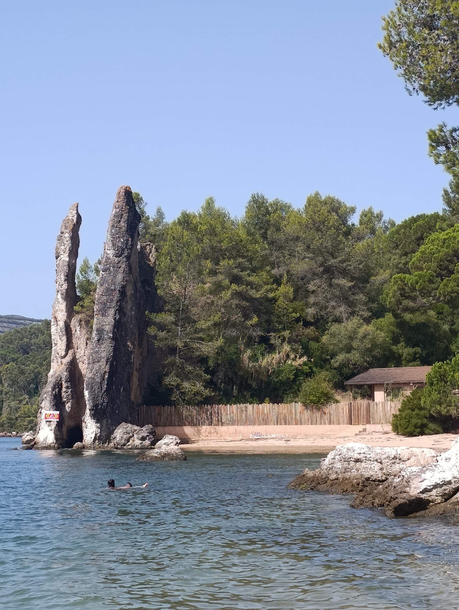 The image depicts a scenic view of the coastline in Setúbal, Portugal. In the foreground, the clear blue water of the sea gently laps against a rocky shore. Two individuals are visible swimming in the water. To the left, a prominent rock formation rises from the sea, characterized by its steep, jagged peaks. A red and yellow warning sign is affixed to the rock face near its base. Behind the rock formation, a sandy beach stretches along the coastline, bordered by a rustic wooden fence. Further inland, a dense forest of green trees forms a verdant backdrop, with a small, brown-roofed building nestled amongst the foliage. The sky above is a clear, cloudless blue.