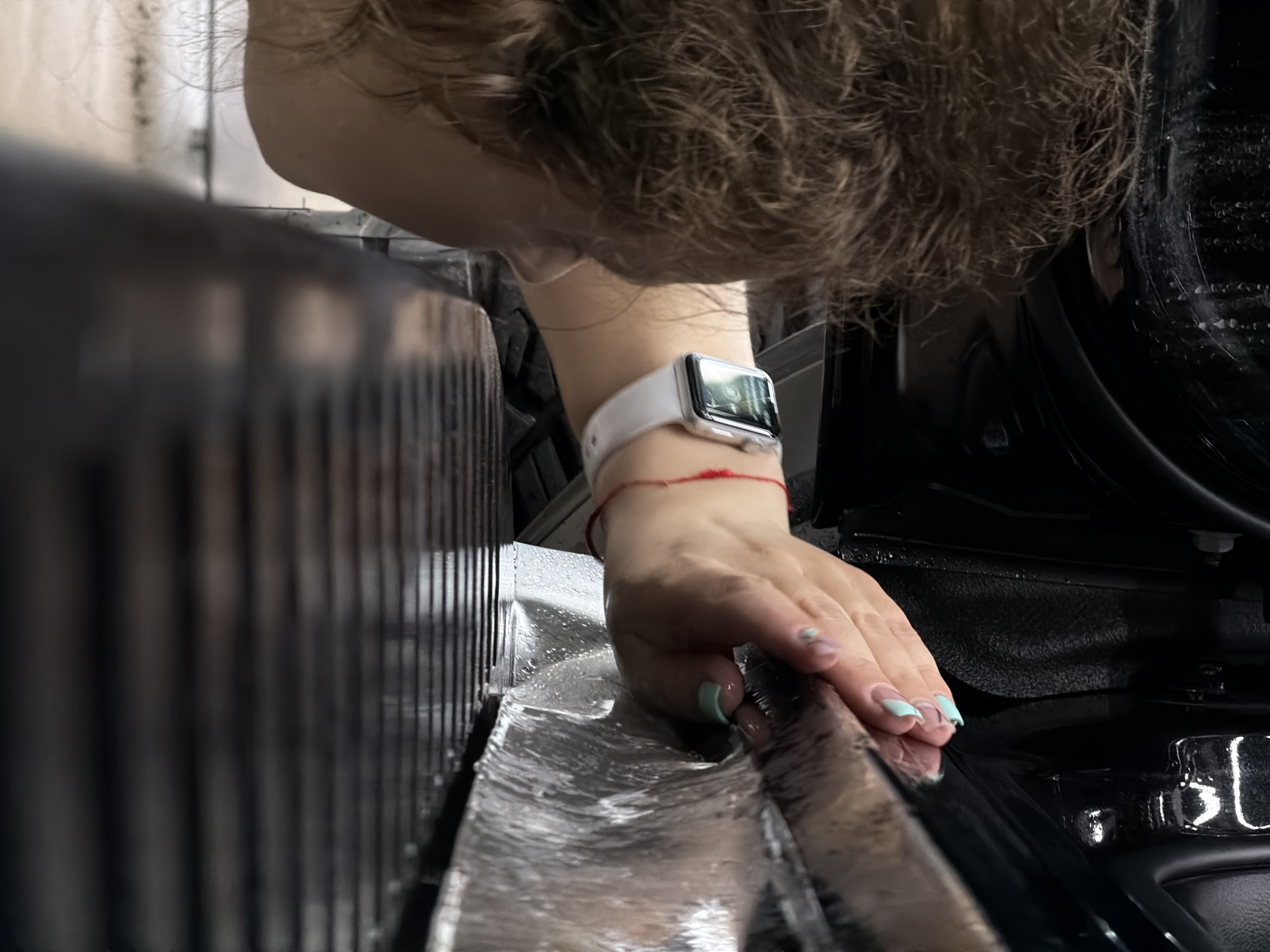 A person with light brown, curly hair is positioned above and facing downward towards the left. The person is wearing a white smartwatch on their left wrist, adorned with a red string bracelet. The person's left hand is on a shiny, reflective surface. The person has painted nails with a light green design. In the background is a dark, vertically ridged structure. The environment appears to be a car cleaning setting. There is no indication of the location being Krasnoyarsk, Russia.