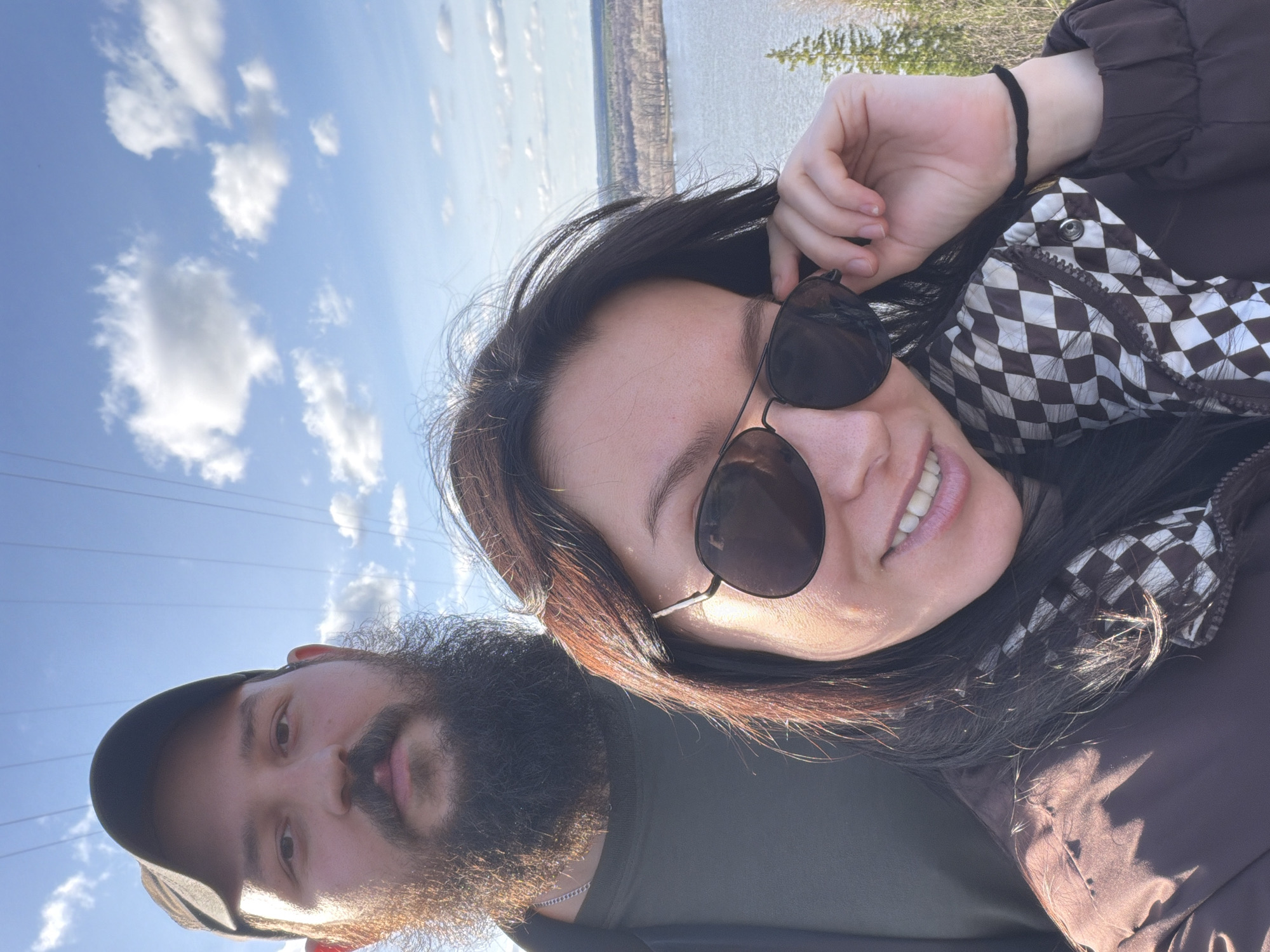 The image shows a selfie taken outdoors, likely during the daytime due to the bright sky with scattered clouds visible in the background. A body of water is seen in the distance.
In the foreground, a woman with dark hair and sunglasses smiles at the camera. She wears a checkered pattern garment under a brown jacket and a black bracelet on her left wrist. Beside her is a man with a thick, full beard and a black cap. He looks directly at the camera.
Overhead power lines are visible in the upper left corner of the frame.
The location appears to be in a natural setting with some vegetation in the background, though the specific features do not confirm the city of Chaykovsky, Russia.