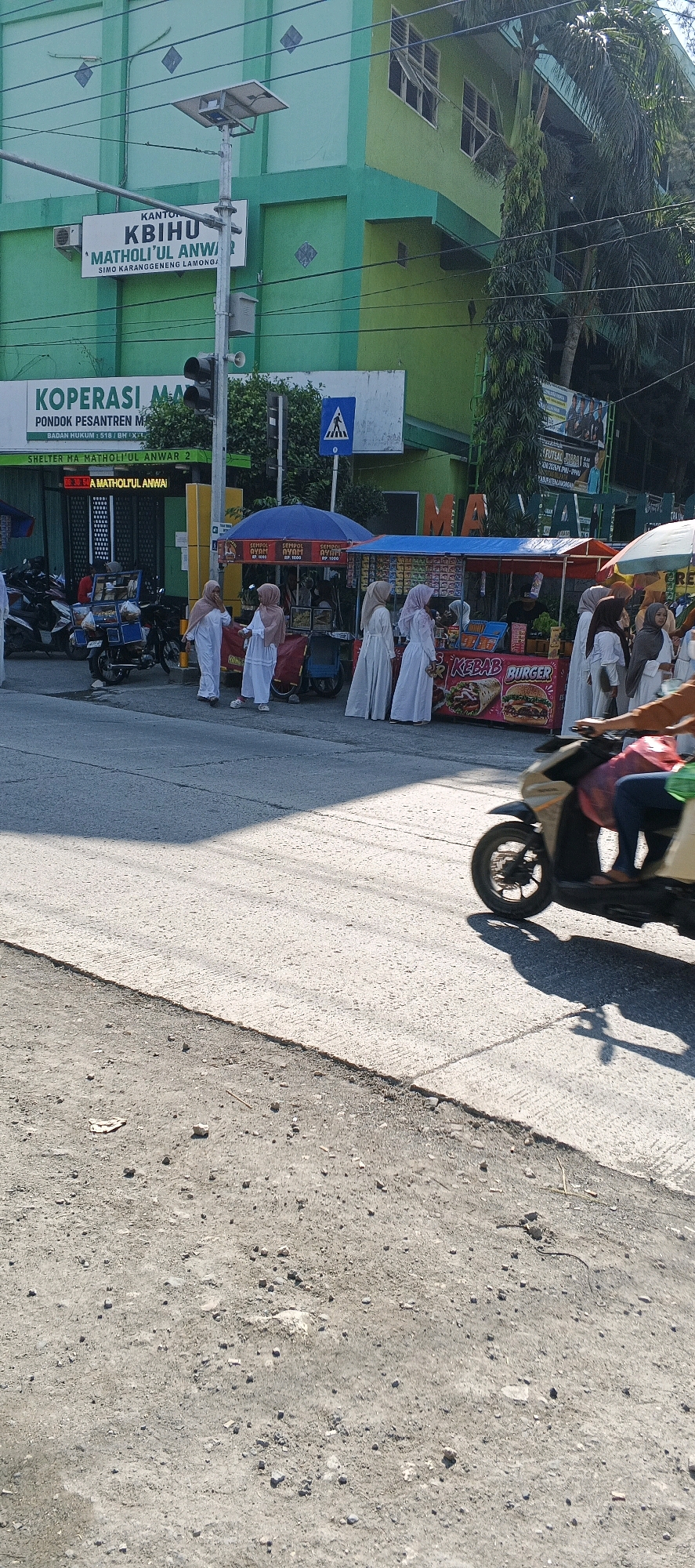 The image depicts a street scene in Sungelebak, Indonesia, in front of a green building with the signage "KANTOR KBIHU 'MATHOLIFUL ANWAR'" and "KOPERASI MA." The building appears to be a school or institution. Several people, mostly women dressed in white Islamic attire, are seen near food stalls set up on the sidewalk. One food stall displays images of "KEBAB" and "BURGER." Motorbikes are parked on the side of the road, and a person on a motorbike is captured in motion in the foreground, casting a shadow on the asphalt. A traffic light and a pedestrian crossing sign are also visible. The environment suggests a sunny day with some shadows cast on the road.