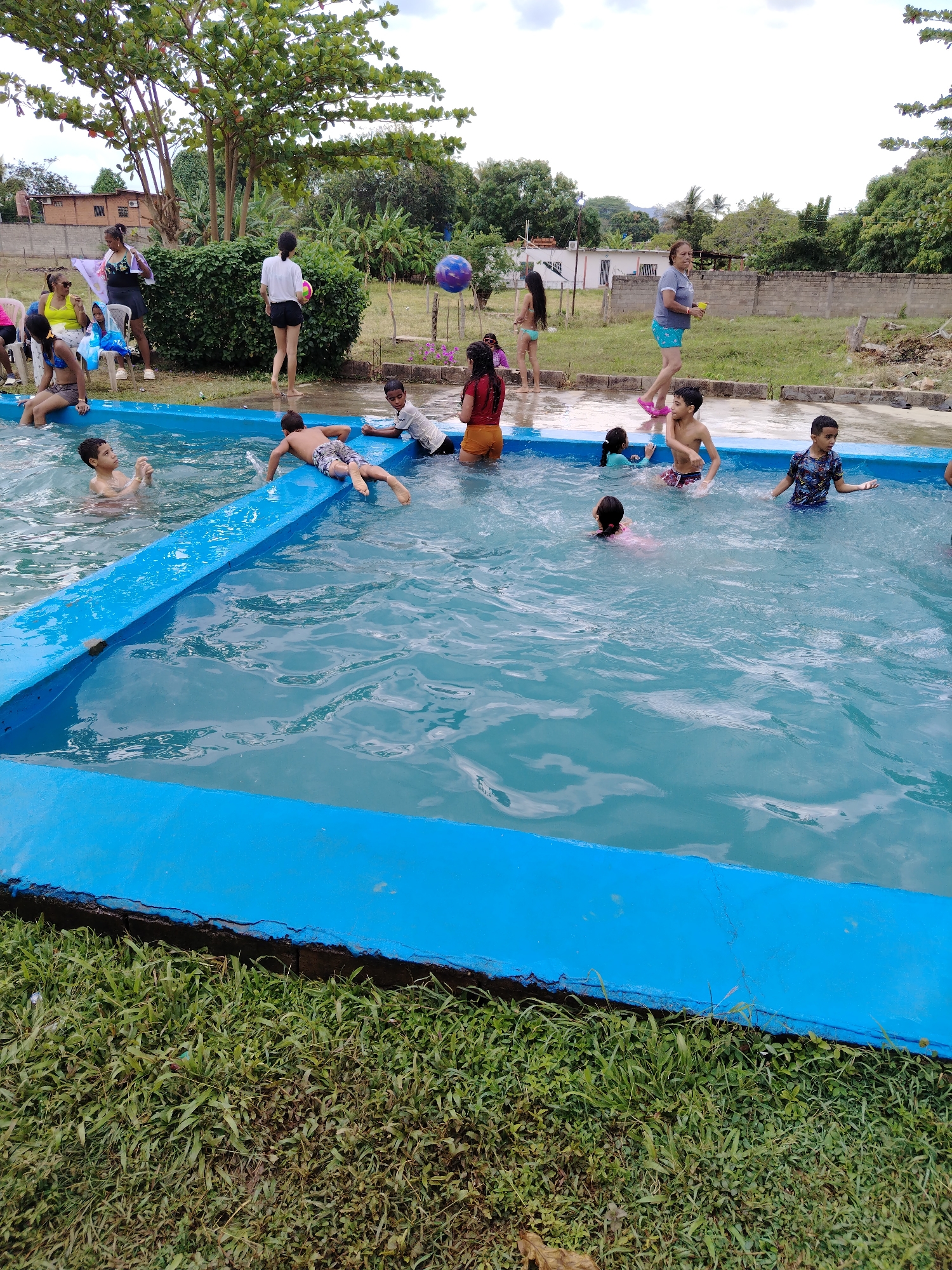 A group of children are playing in a rectangular, blue-rimmed swimming pool. Some are swimming, others are sitting or lying on the edge of the pool. Several adults are standing or sitting nearby. The pool is surrounded by grass and vegetation. In the background, there are houses and a wall. The location is identified as the city of Tinaquillo, Venezuela.