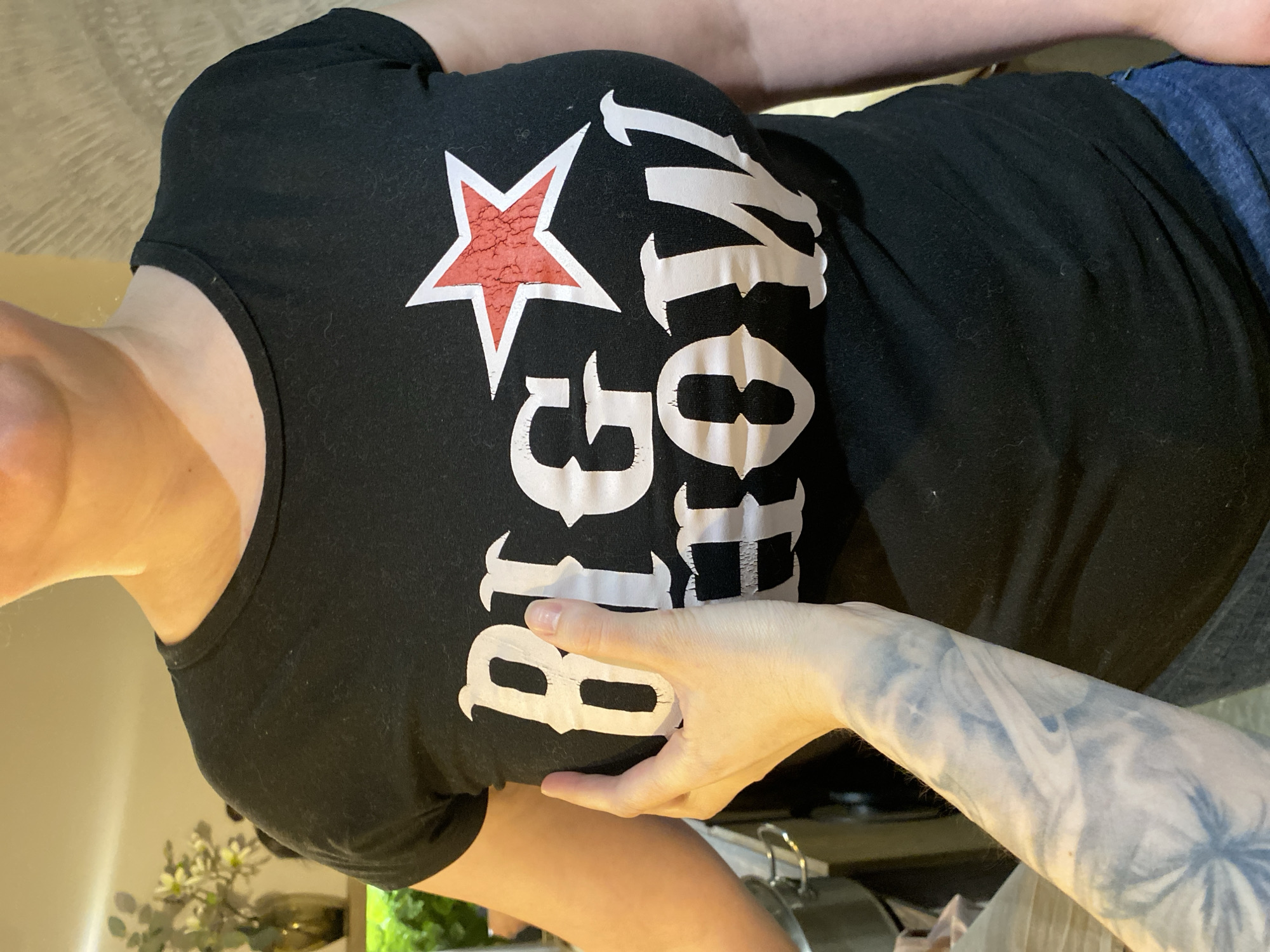 A person is wearing a black t-shirt with a white star and lettering reading "Big Momma" in a room with beige walls and some greenery. A tattooed arm is visible to the right, with a hand partially obscuring the shirt. The exact location is not identifiable as being in Votkinsk, Russia, nor can the environment be more precisely characterized from the given image.