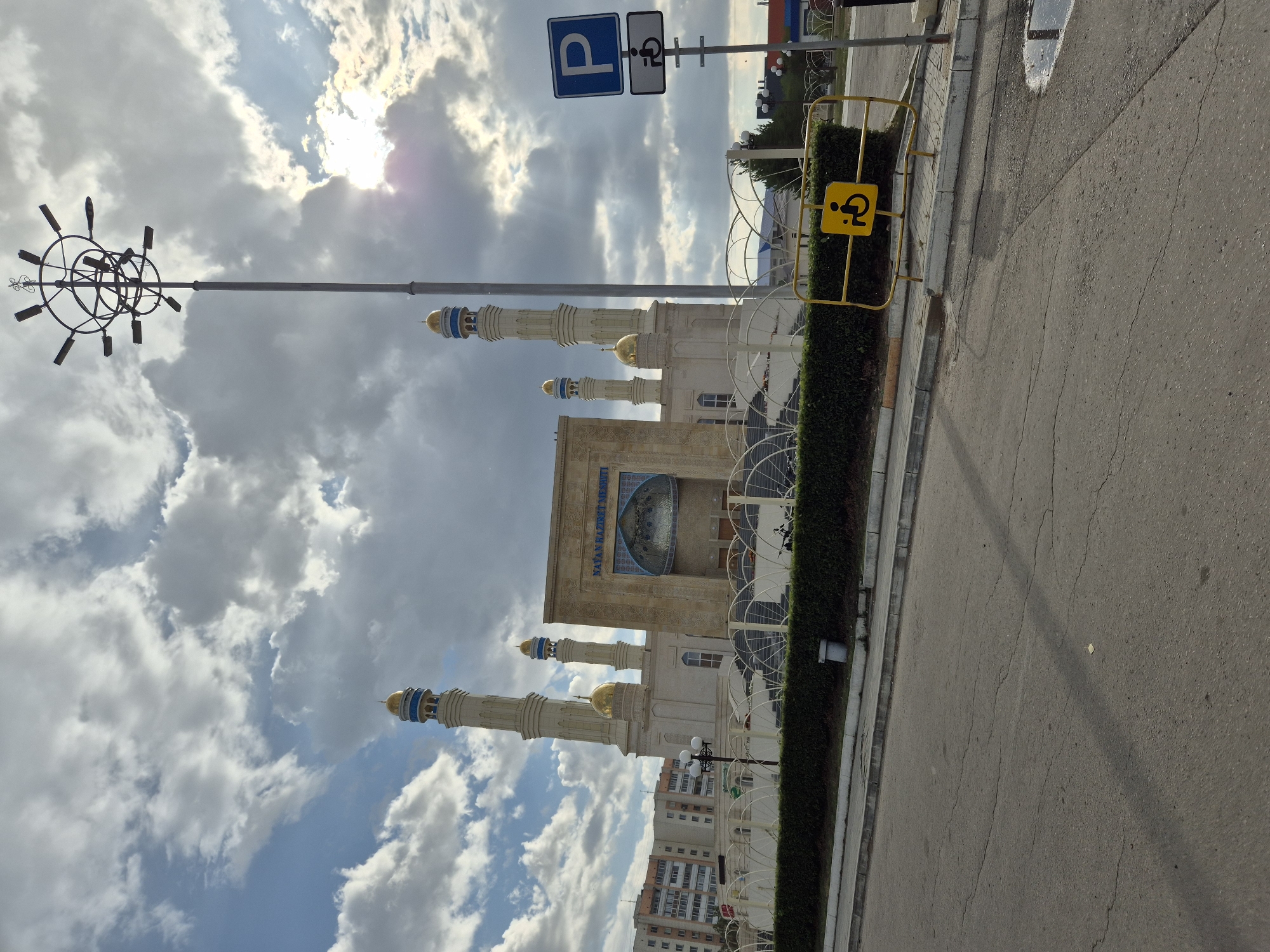 The image shows the Nayman Hazrat Mosque in Kokshetau, Kazakhstan, on a cloudy day with the sun peeking through the clouds. The mosque is a light-colored building with gold-domed minarets. In front of the mosque, there is a decorative white fence and a hedge. A tall lamppost with multiple lights stands to the right of the mosque, and further to the right, there are parking signs, including a blue P sign and a sign indicating a parking spot for people with disabilities. The foreground shows a cracked asphalt road and a sidewalk. In the background, there are apartment buildings. No people are visible in the image.