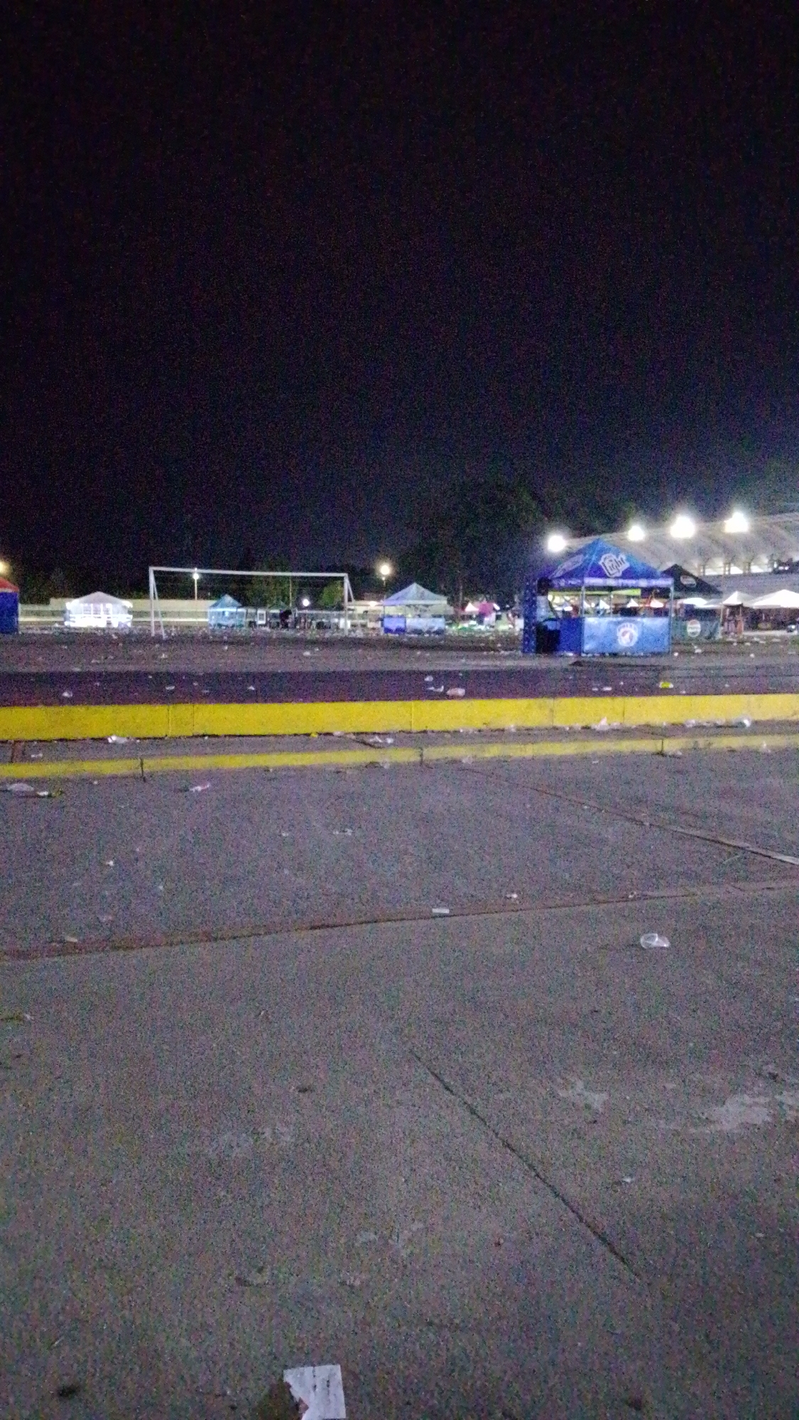 The image depicts an outdoor area at night, possibly a parking lot or open space in Tinaquillo, Venezuela. In the background, there are tents or stalls, some with visible branding like "Miller Lite." A soccer goal is also visible. Debris and litter are scattered across the ground. The area appears illuminated by stadium lights and street lamps. The foreground is a paved surface with cracks and visible trash. No people are distinctly identifiable in the image.