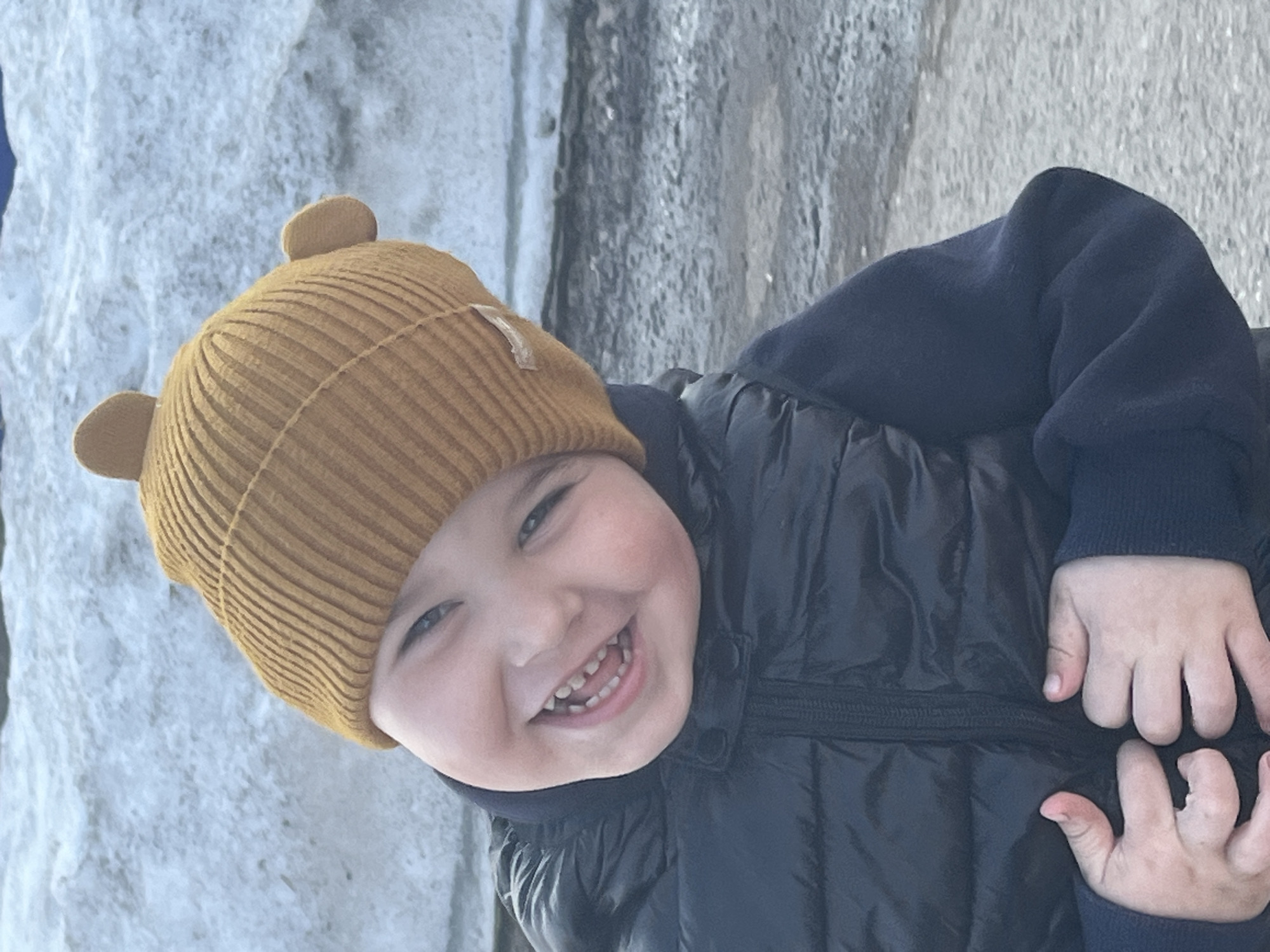 A young child, likely a toddler or preschooler, is smiling broadly at the camera. The child is wearing a mustard-yellow knit hat with small ears and a dark-colored, possibly navy blue, puffer jacket with a matching long-sleeved shirt underneath. The child's hands are visible, resting on the jacket. The background appears to be a textured concrete or stone surface. No definitive indications point to the city of Павловка, Russia.