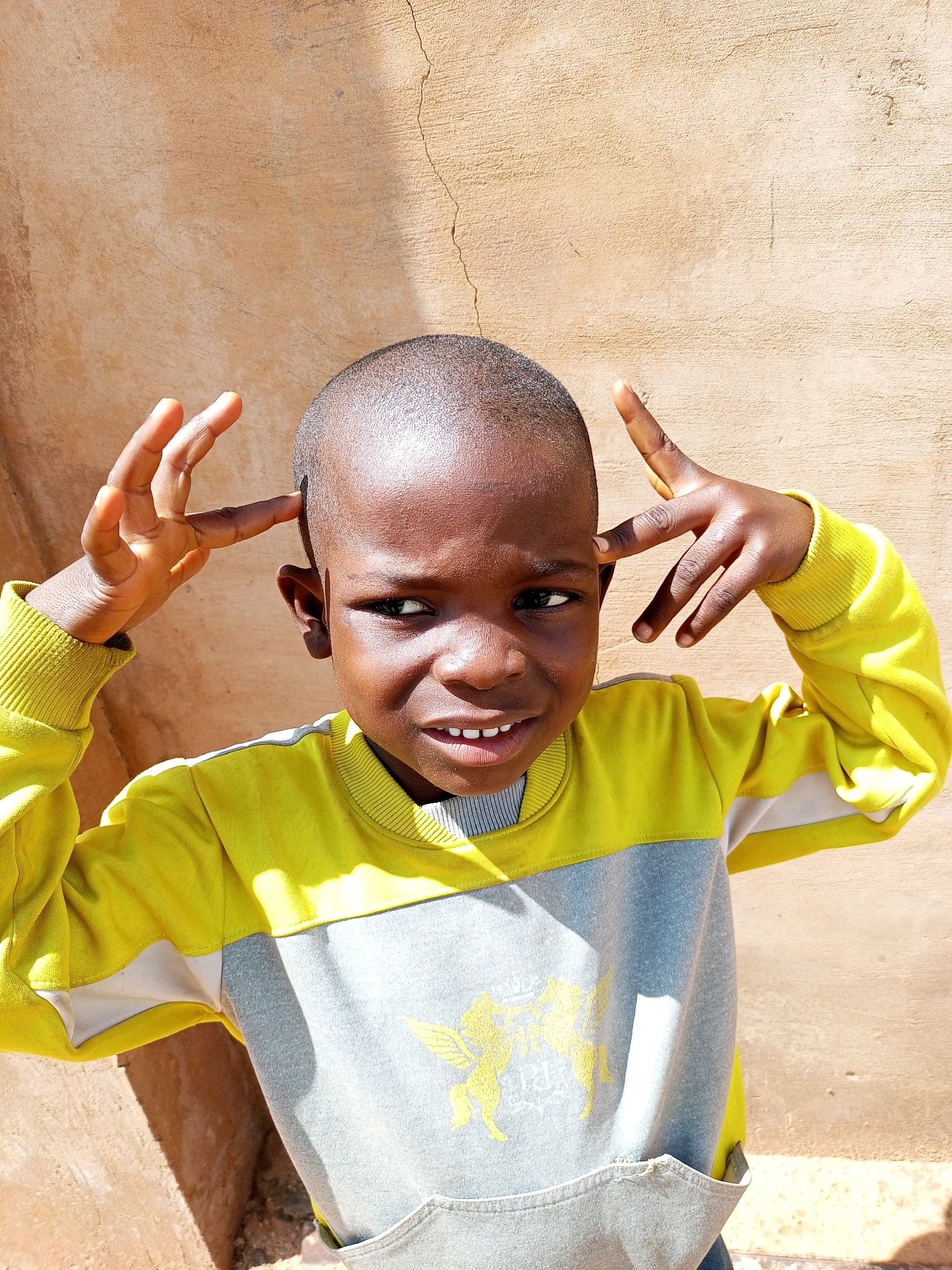A young boy of African descent, with dark skin and short, dark hair, is pictured outdoors. He is wearing a yellow and gray long-sleeved shirt with a yellow graphic design on the front. The boy is looking slightly to his left with his mouth open, revealing his teeth. His left hand is raised to the side of his head, with fingers splayed and bent, touching his temple. His right hand is also raised to the side of his head, with two fingers extended and touching his temple. He is standing in front of a rough, tan-colored wall that appears to be made of stucco or concrete. Sunlight casts shadows on the wall and the boy's clothing. The background suggests an outdoor environment, possibly in a rural or semi-urban setting within Tempure, Nigeria.