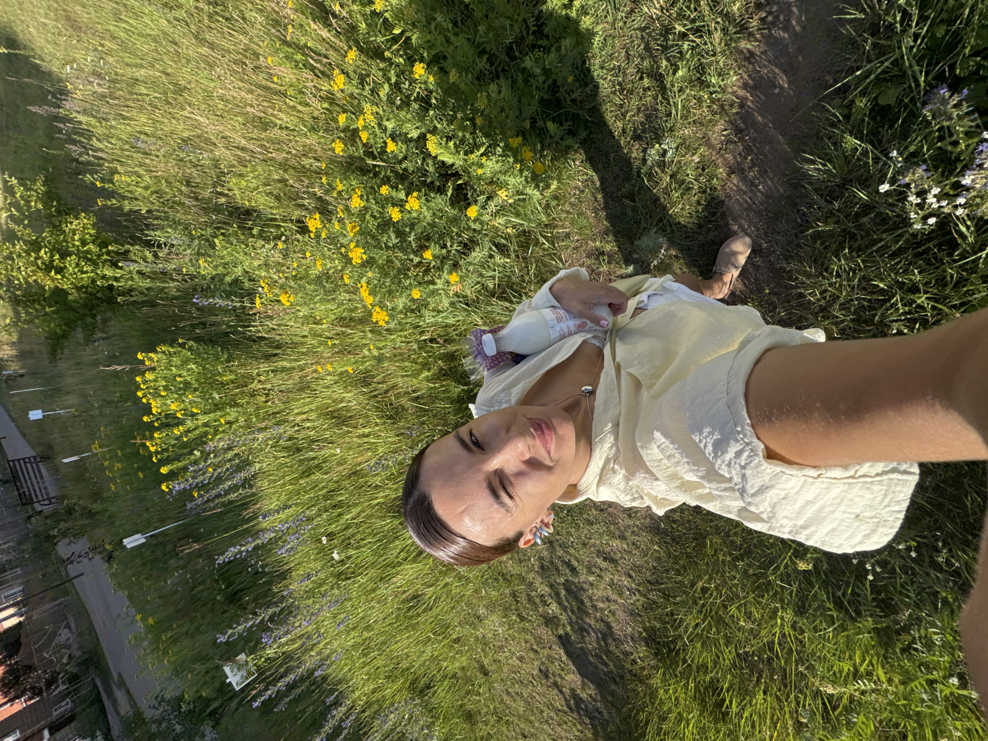 In a sunlit, grassy area, a woman is taking a selfie. She is wearing a light yellow, loose-fitting garment and is holding a clear plastic bottle filled with a white liquid. Her left eye is closed in a wink. The background is filled with tall, green grass and scattered yellow and purple wildflowers, suggesting a rural or park-like setting. The image was taken in the city of Chaikovsky, Russia, likely during a warm day due to the bright sunlight and the woman's attire.
