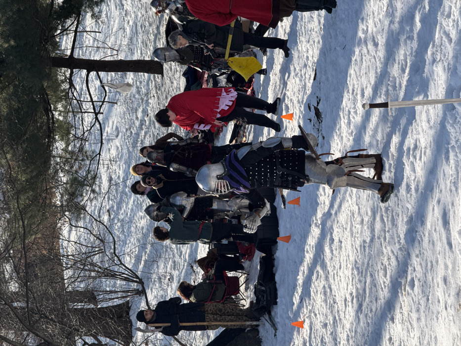 A group of people dressed in medieval armor stand in a snowy field. Some are wearing red and black tunics, while others are fully armored. A few orange cones are scattered around. Trees are visible on the left side of the frame. A sword lies on the snow-covered ground to the right.